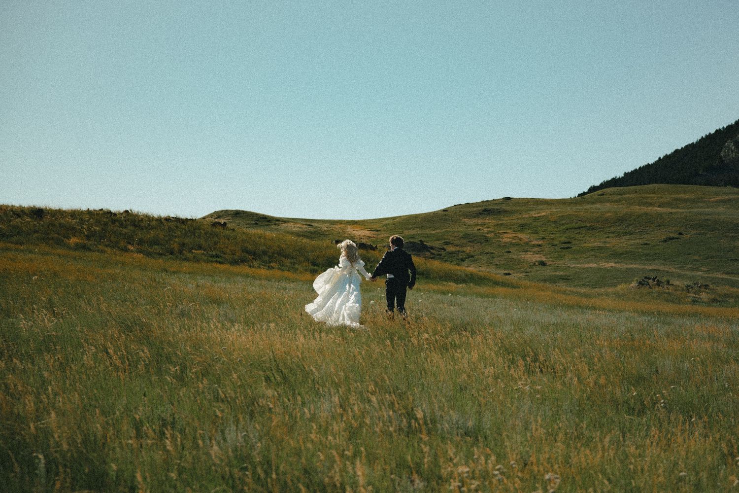 A bride in a white gown and groom run hand in hand through a lush green meadow on their wedding day.