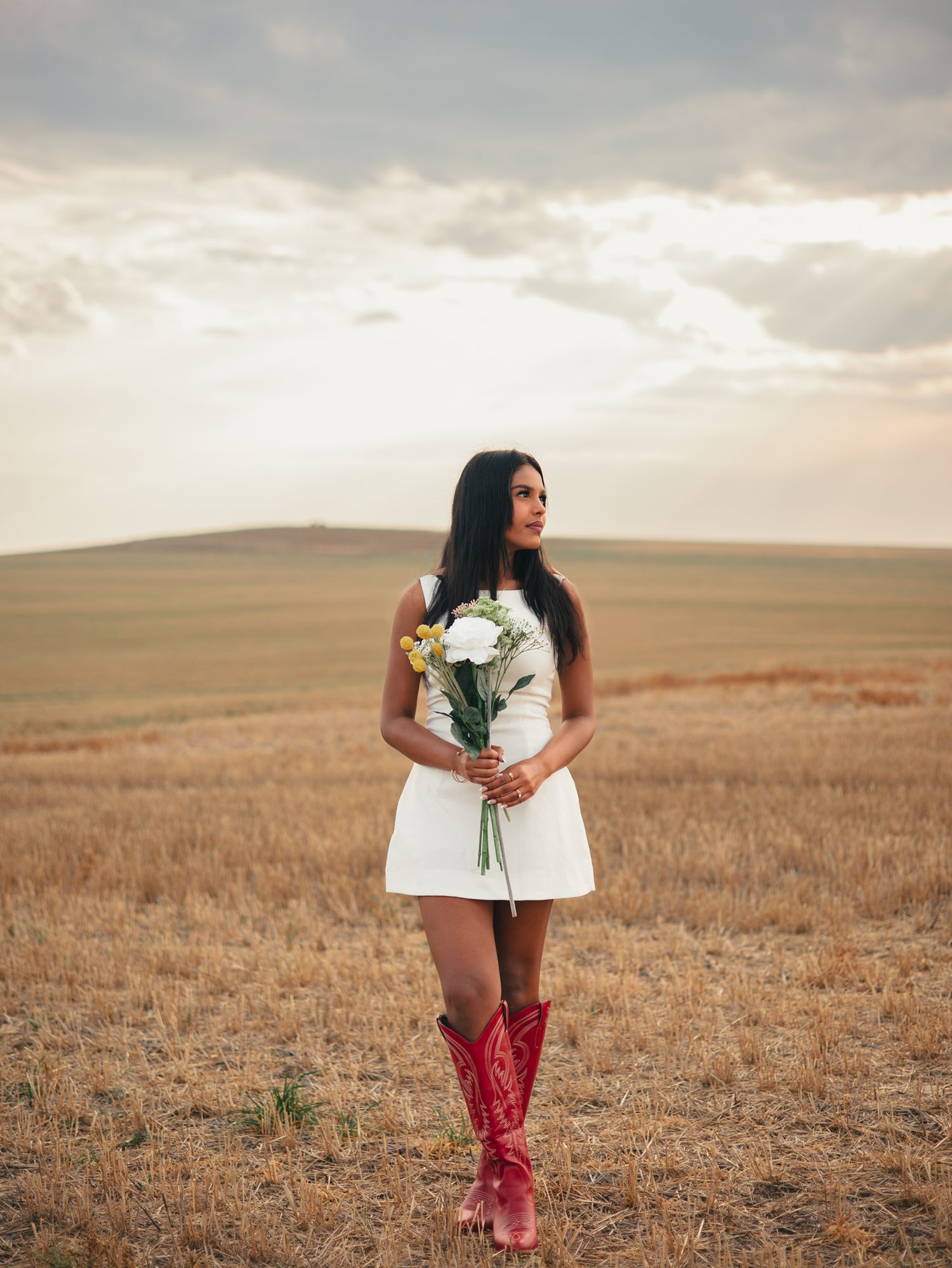 A figure in a white dress and red pants stands in a golden field at sunset holding flowers. photo, Great Falls, MT