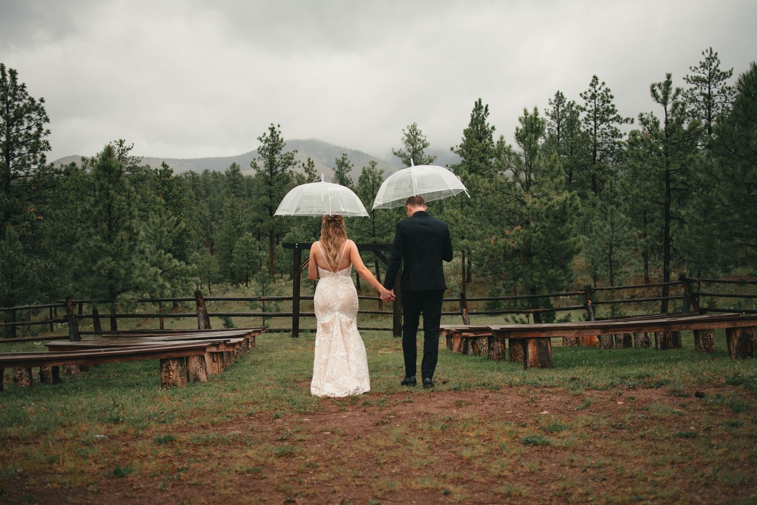 Bride and groom walking hand in hand under white umbrellas in a rainy outdoor wedding venue surrounded by pine trees.
