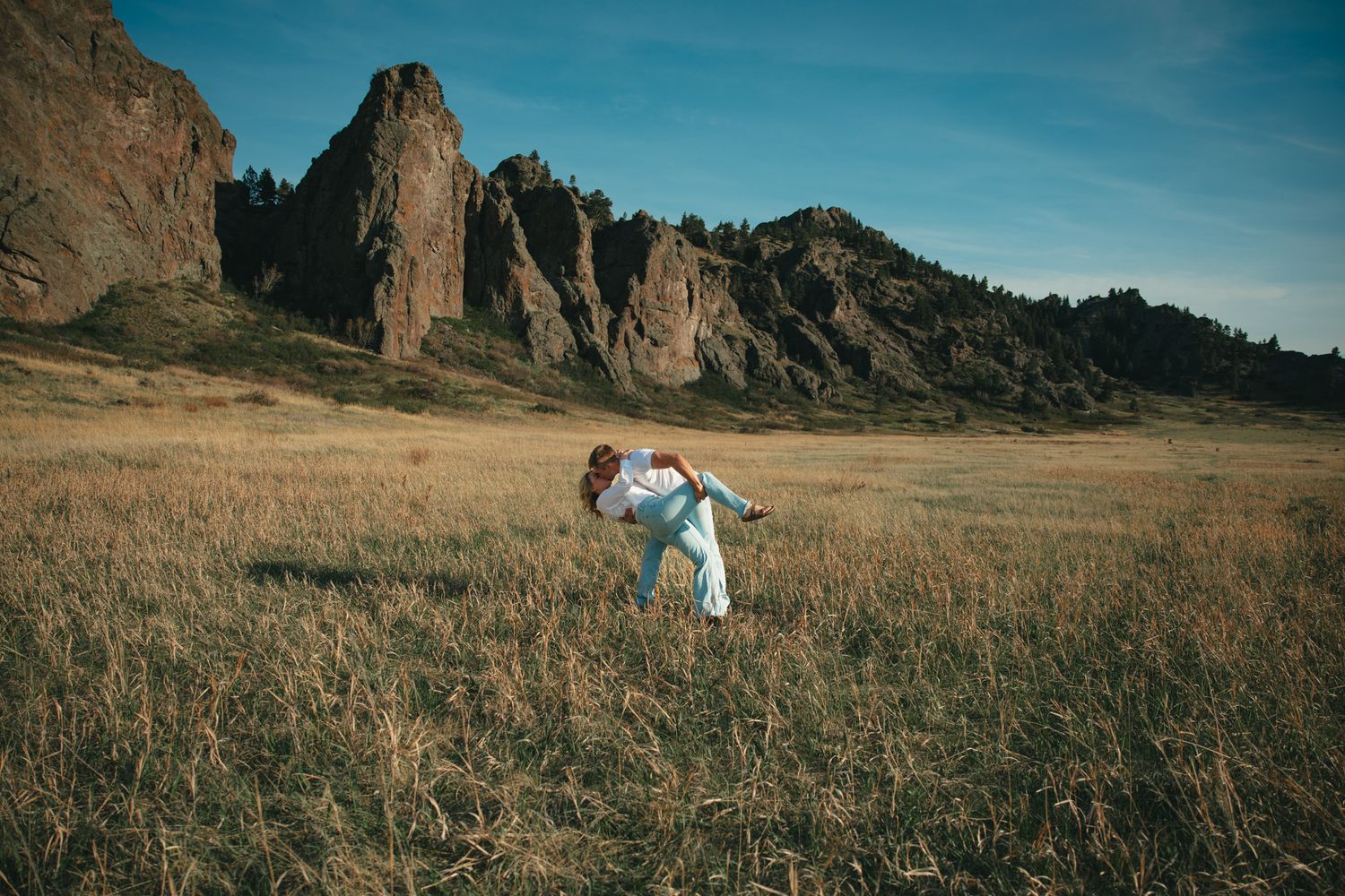 Couple embracing in golden grassland field with dramatic rocky mountain formations under blue sky.