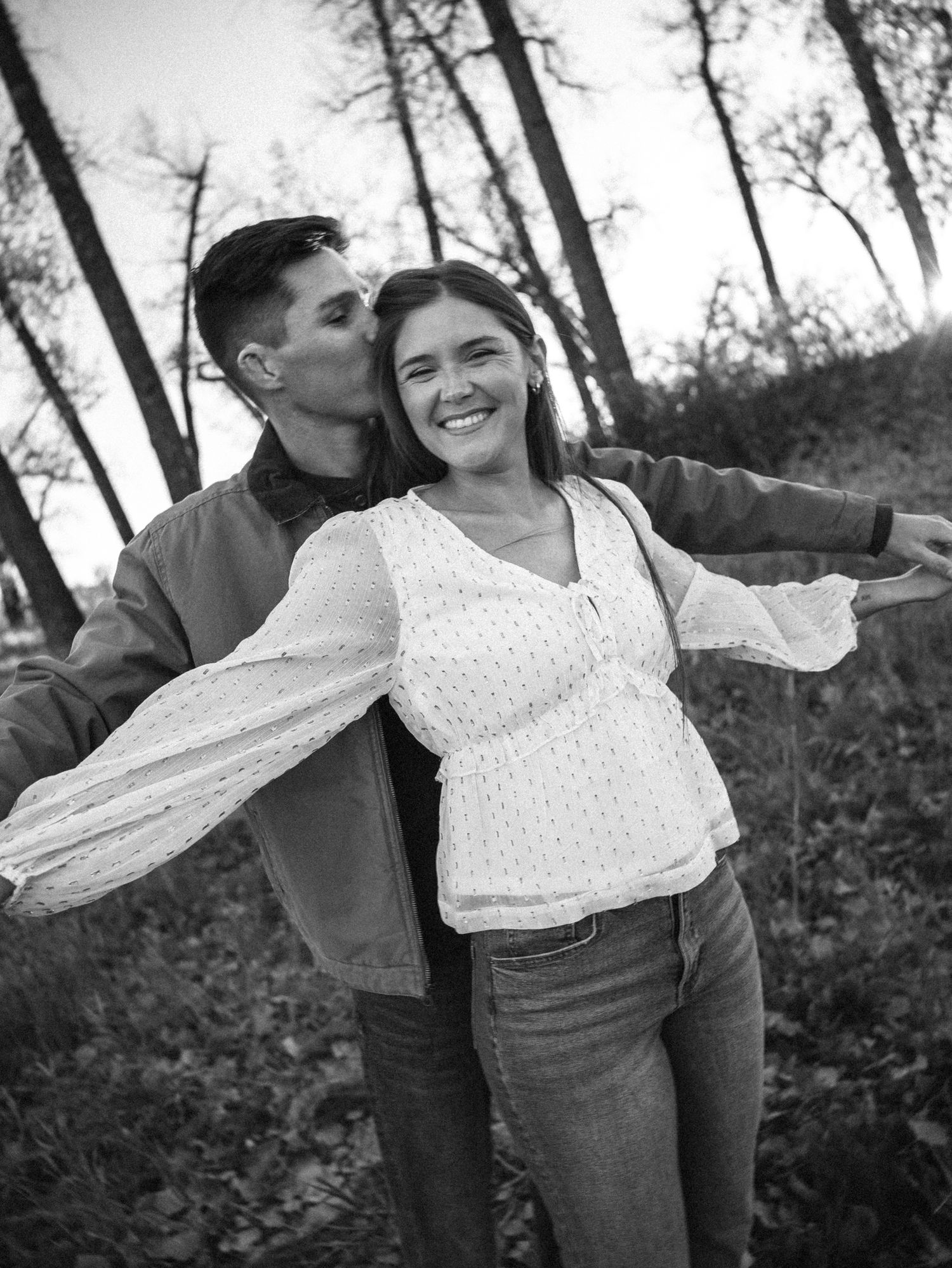 Black and white photo of a smiling couple outdoors, woman with arms outstretched as man kisses her cheek.
