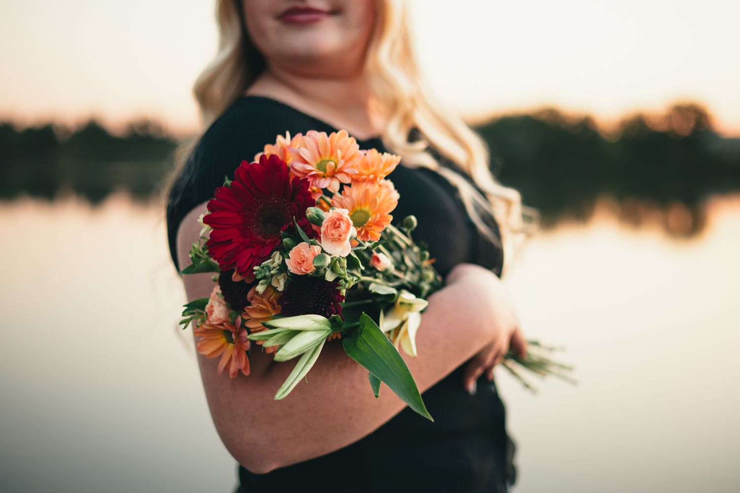 A senior in black holds a vibrant bouquet of orange and pink flowers against a sunset waterfront backdrop. photo, Great Falls, MT