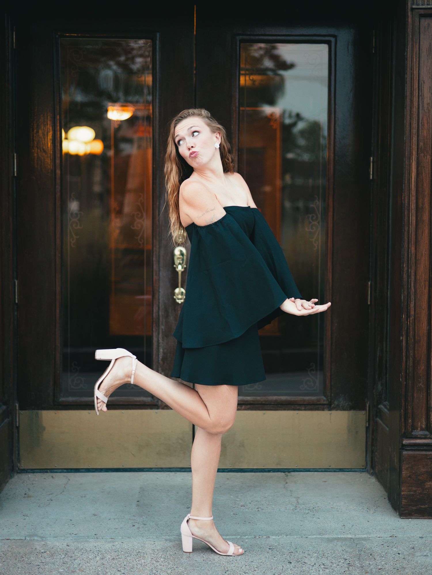 Woman in black dress and silver heels poses playfully in front of dark glass doors at dusk. photo, Great Falls, MT