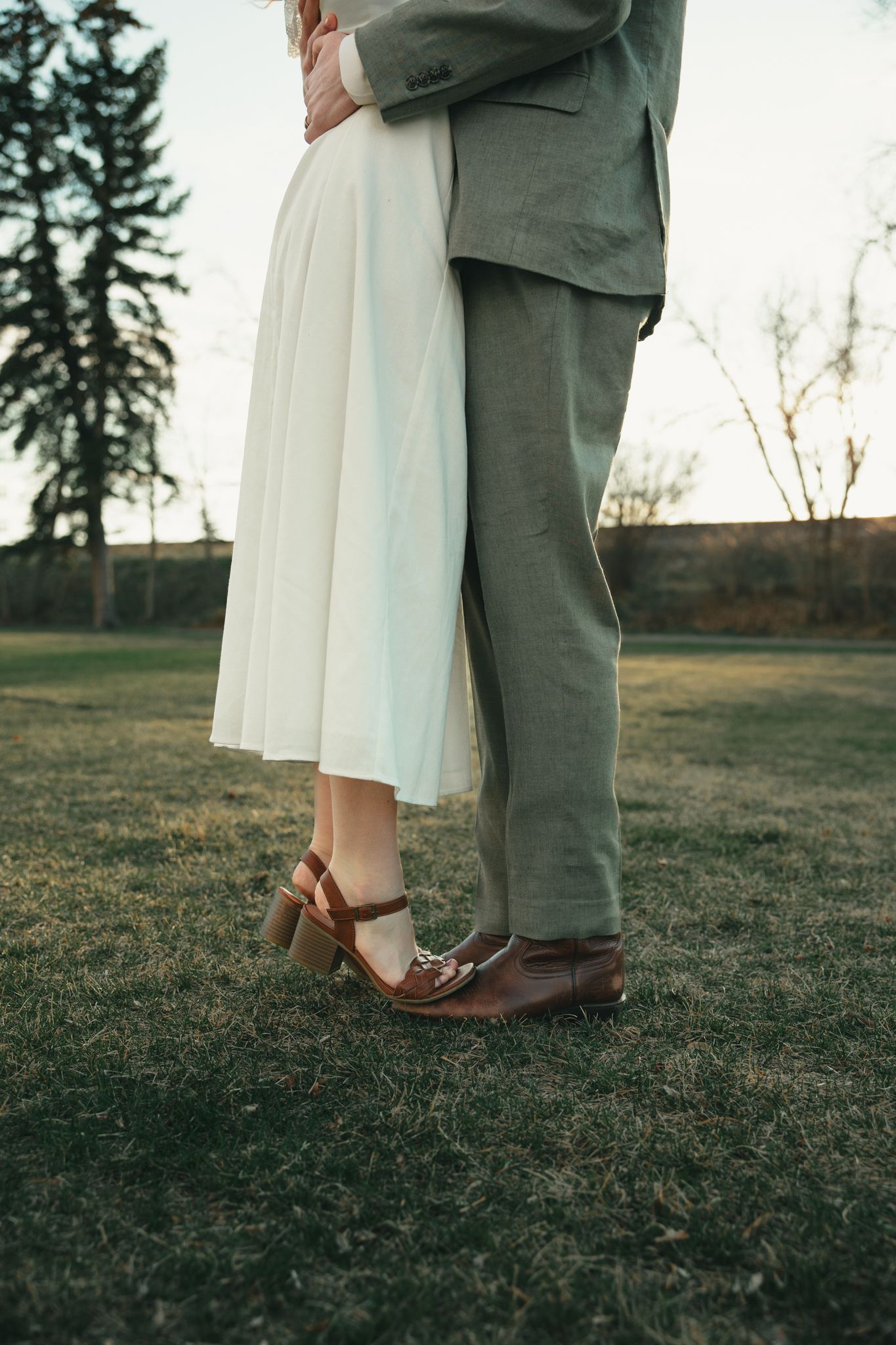 Bride and groom standing close together on green grass, wearing white dress and gray suit with brown leather shoes.