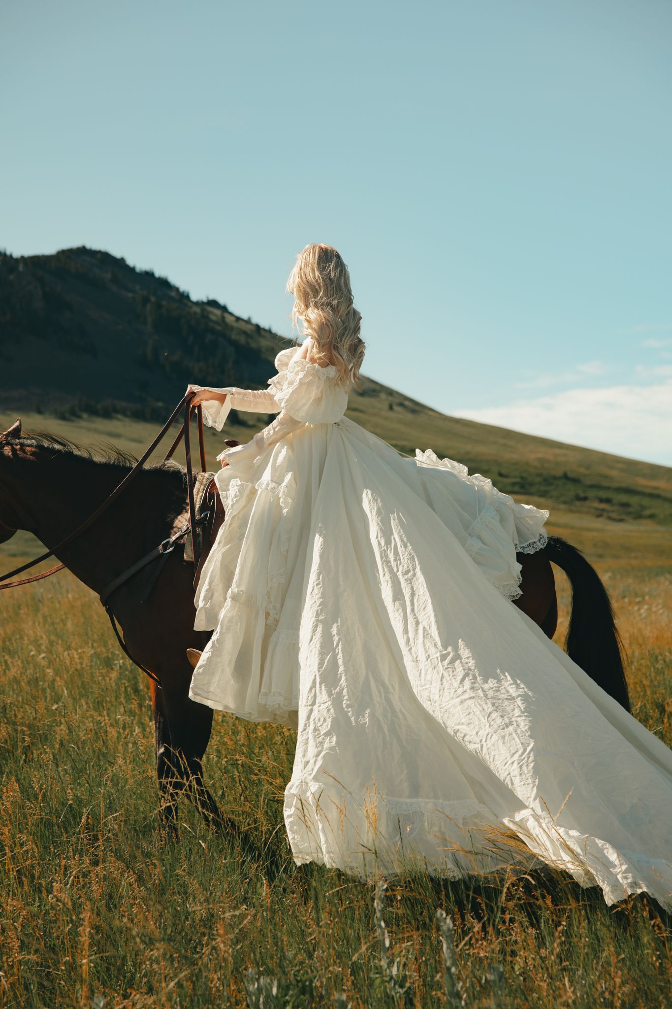 A blonde woman in a flowing white wedding gown rides a dark horse through a scenic mountain meadow.