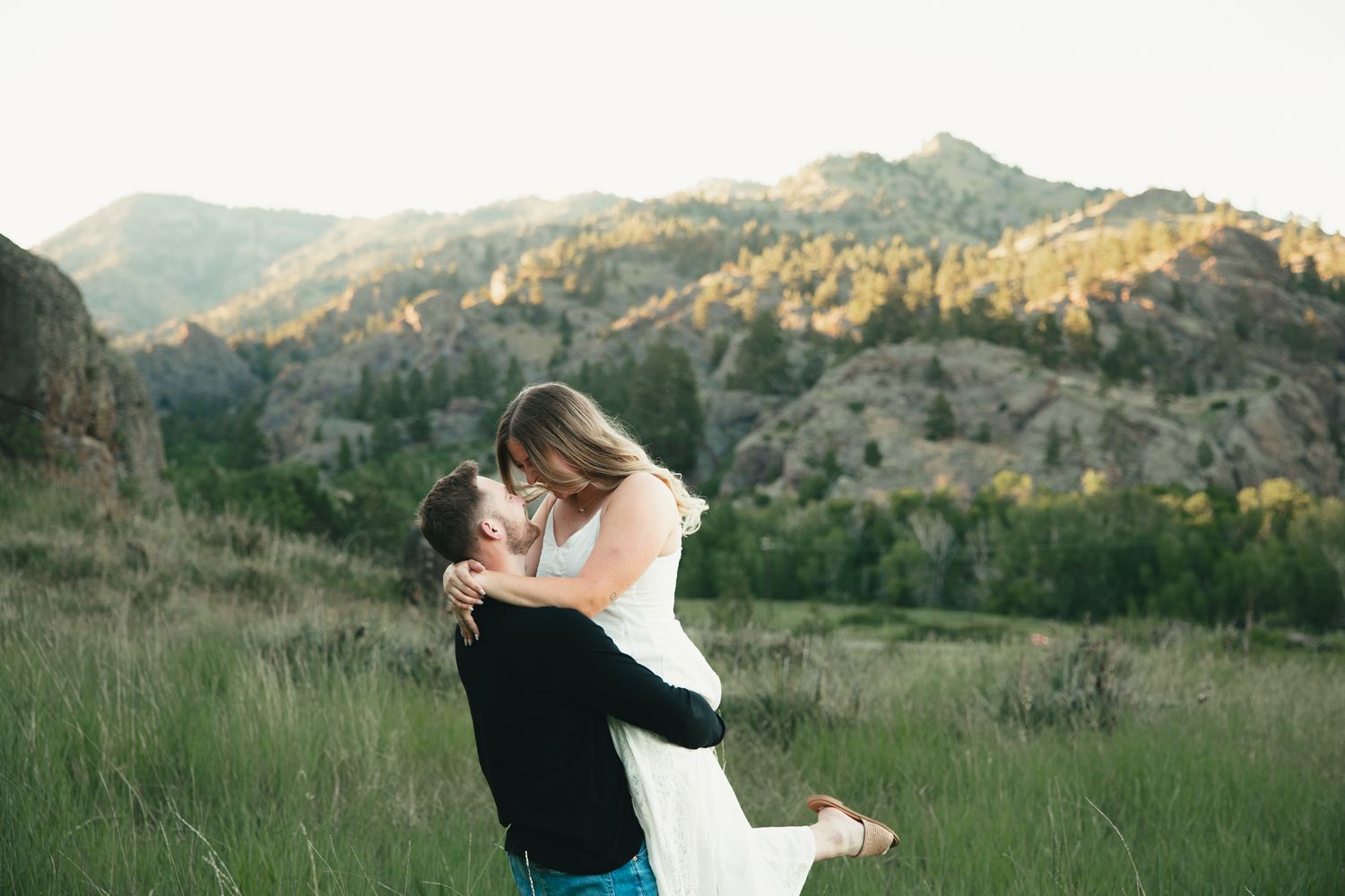 Couple sharing a romantic kiss in a lush green meadow with stunning mountain backdrop at golden hour.