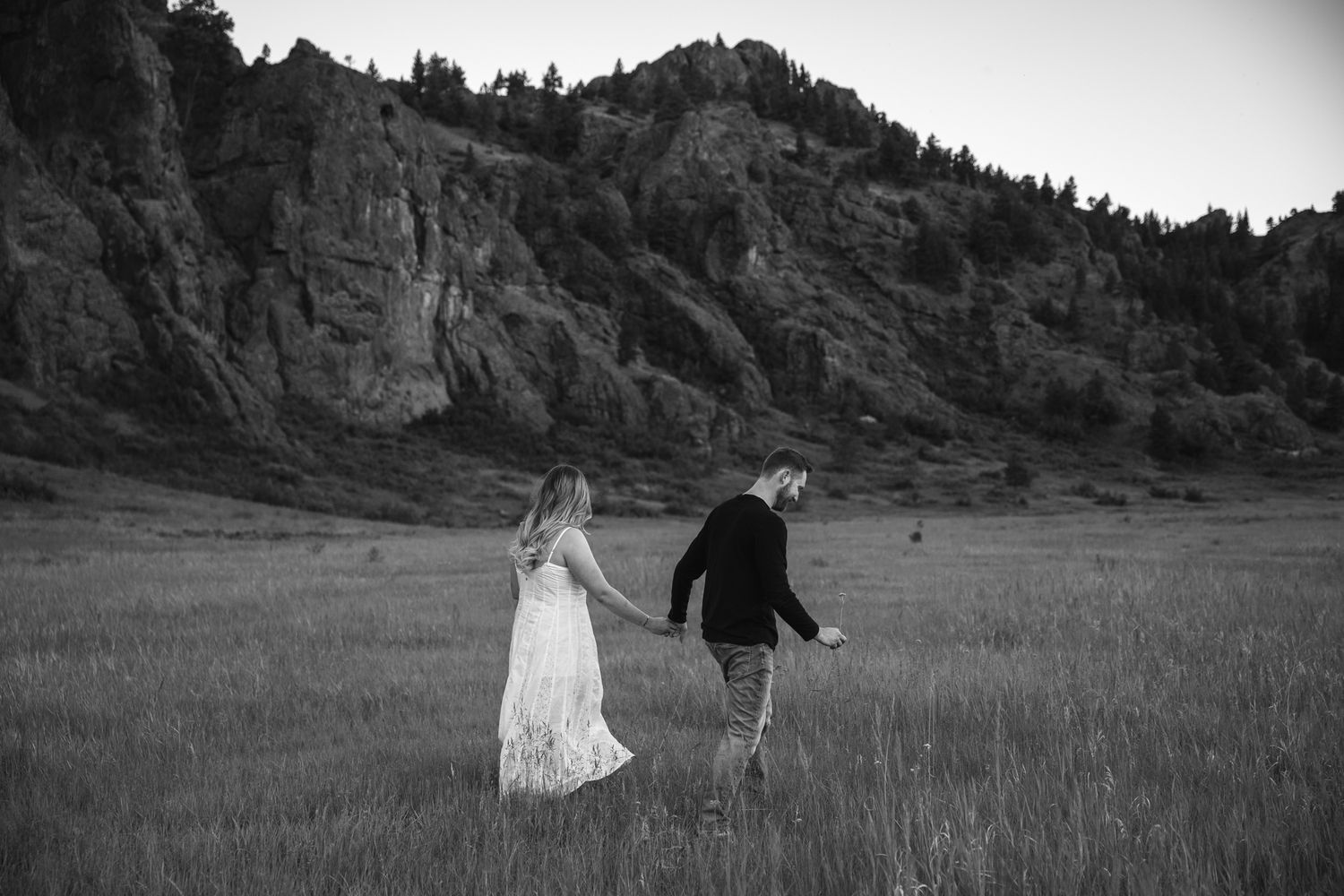 Black and white photo of a couple holding hands walking through a grassy field with rocky mountains behind them.