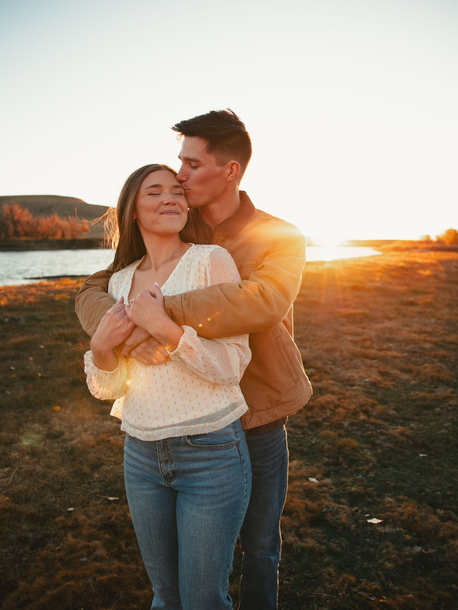 Couple embracing at golden hour sunset by a lake, man kissing woman on cheek while she smiles with eyes closed.