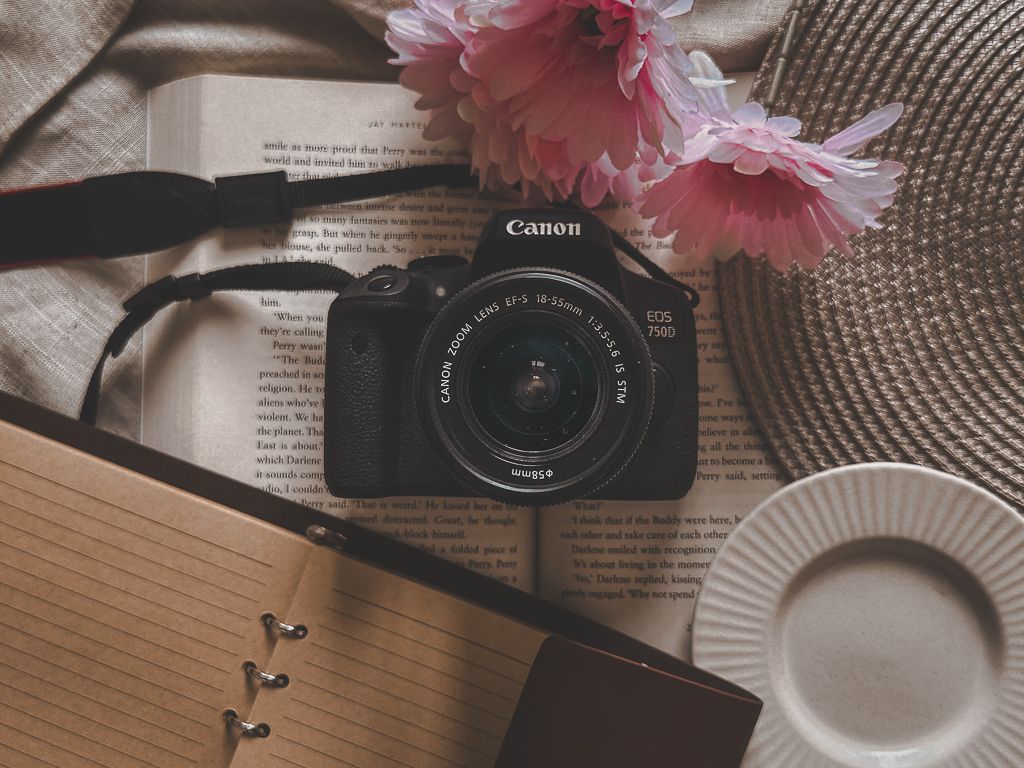 Artistic flatlay featuring pink flowers, a Canon camera, and paper items on textured fabric.
