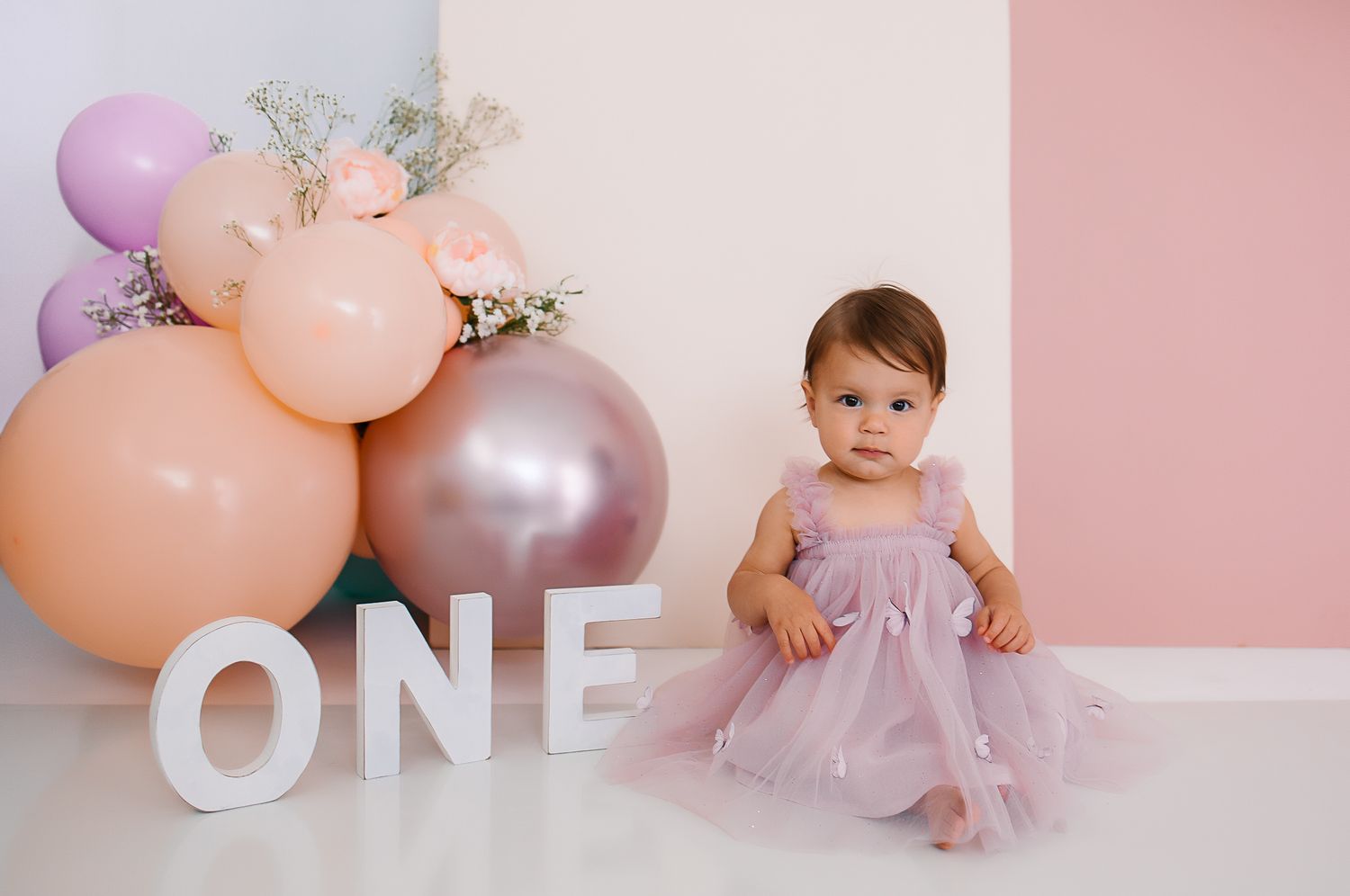 Baby surrounded by pink and peach balloons in tulle dress for first birthday milestone photoshoot.