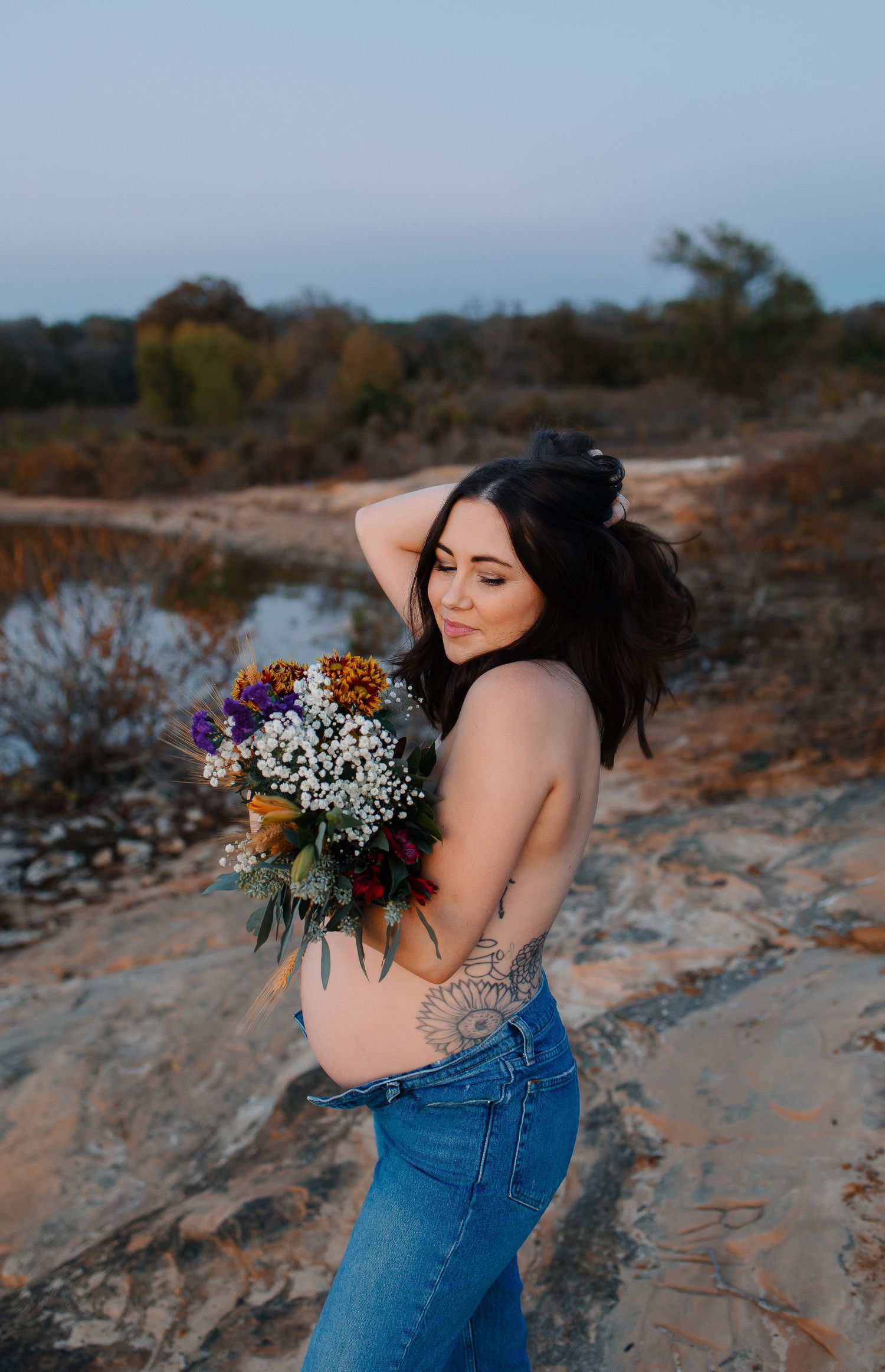 A mother embraces her belly in denim jeans holding a vibrant wildflower bouquet against her stomach during sunset at the lake.