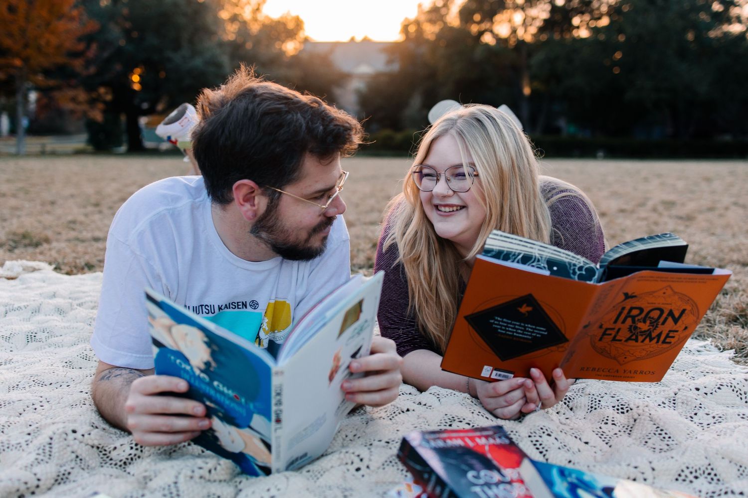 A newly engaged couple lying on a blanket reading books together outdoors at sunset during autumn.