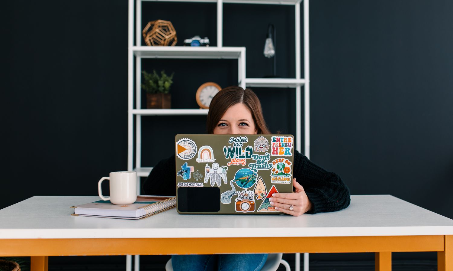 Modern home office setup with white desk, laptop, coffee mug and shelving unit against dark blue wall.