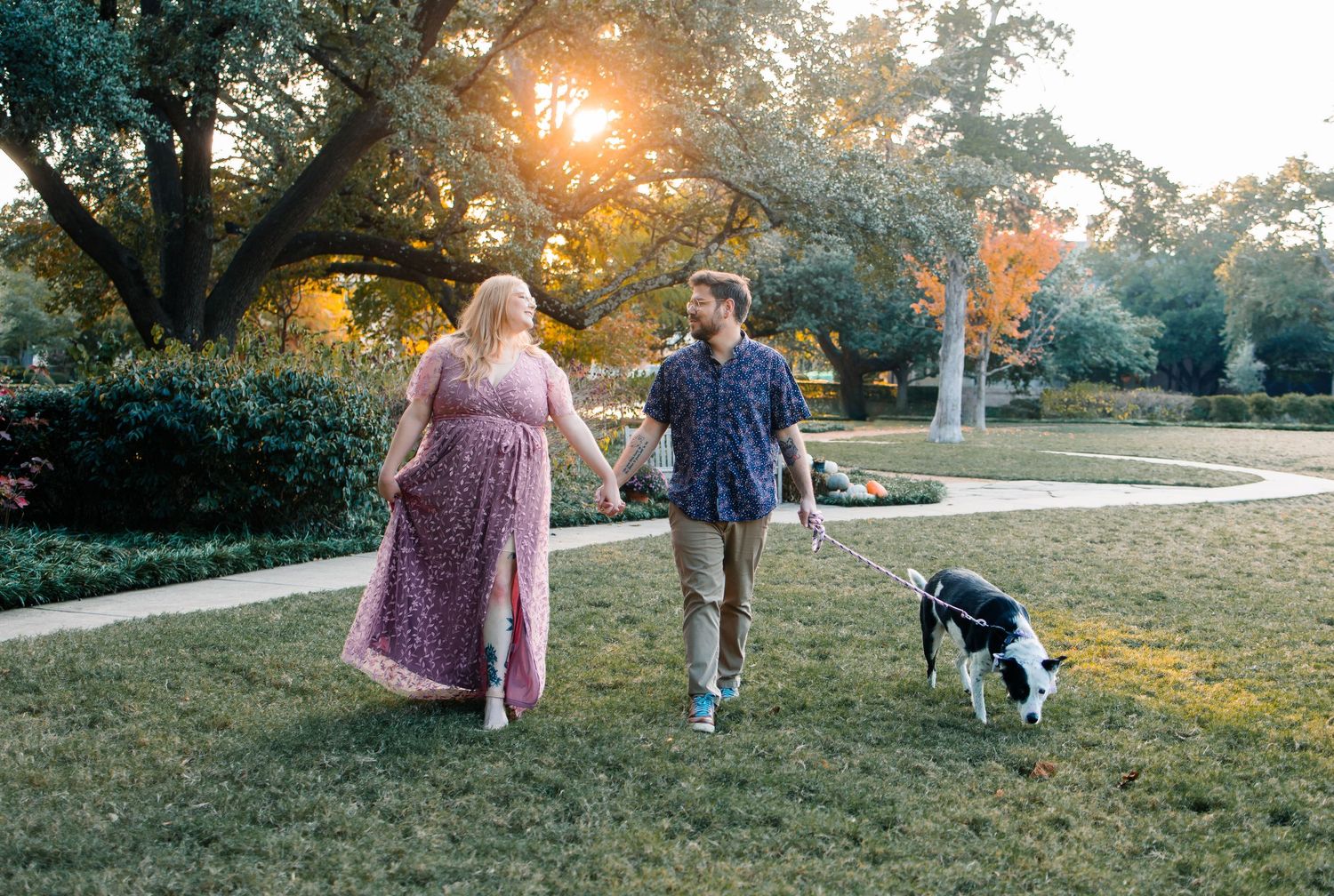 A newly engaged couple walks their dog through a grassy hillside park at sunset, creating a peaceful autumn scene.