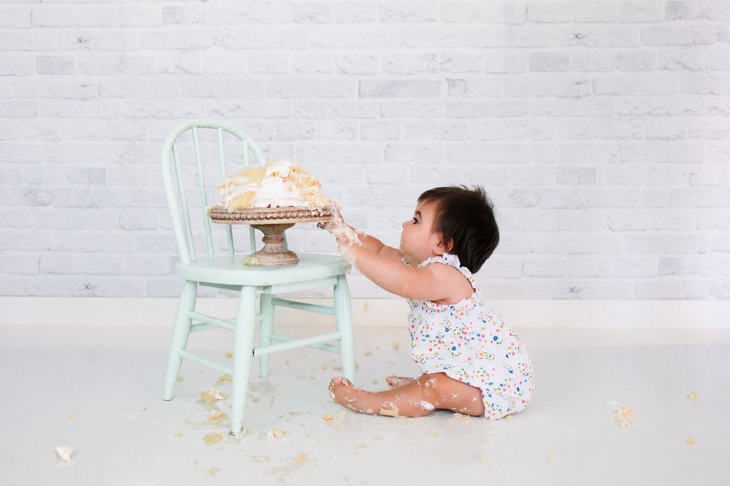 Baby in white wearing floral reaches up to touch cake on mint green chair against light gray brick background.