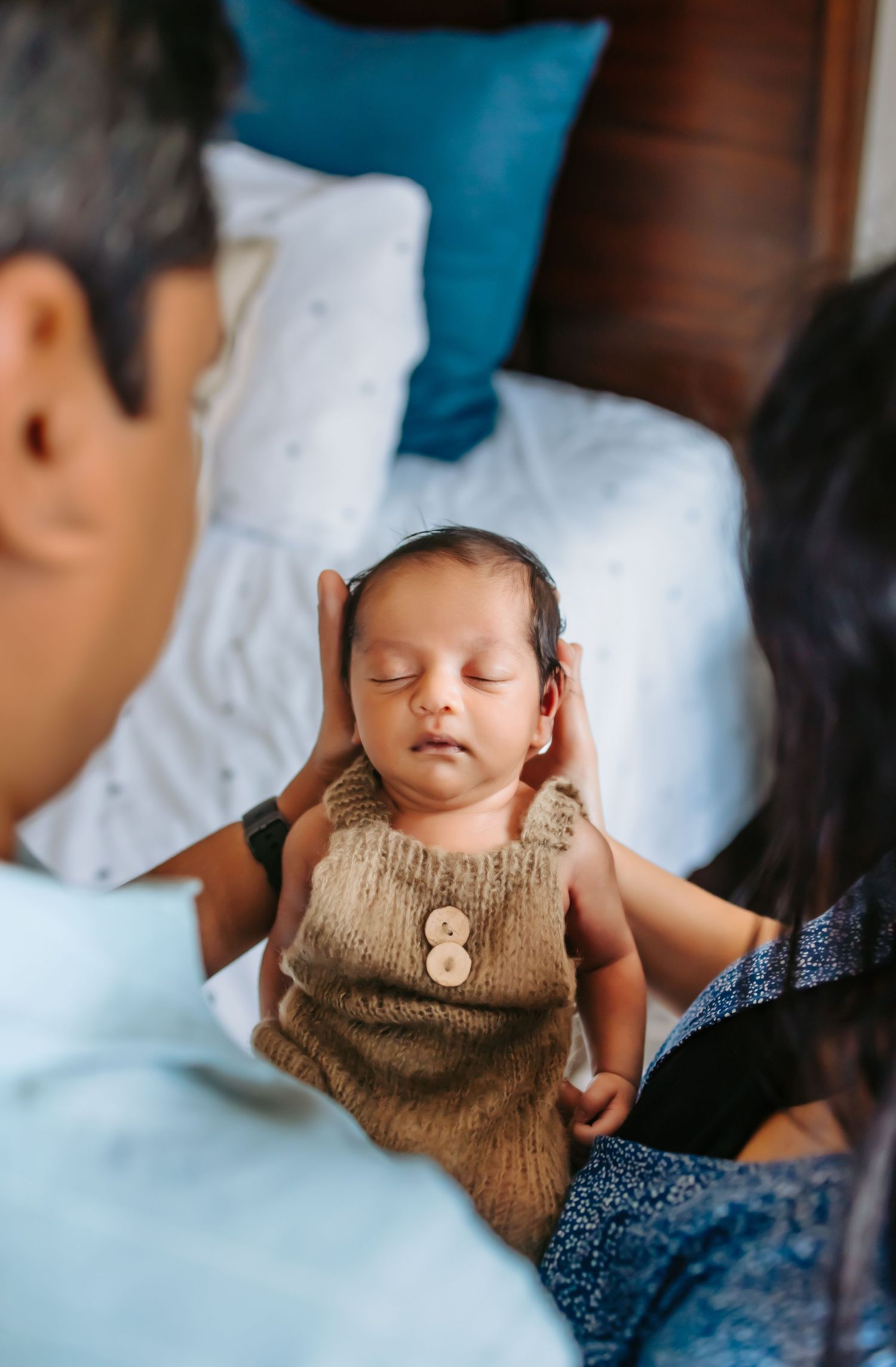 A sleeping newborn baby wearing a brown knitted vest lies peacefully between his mother and father.
