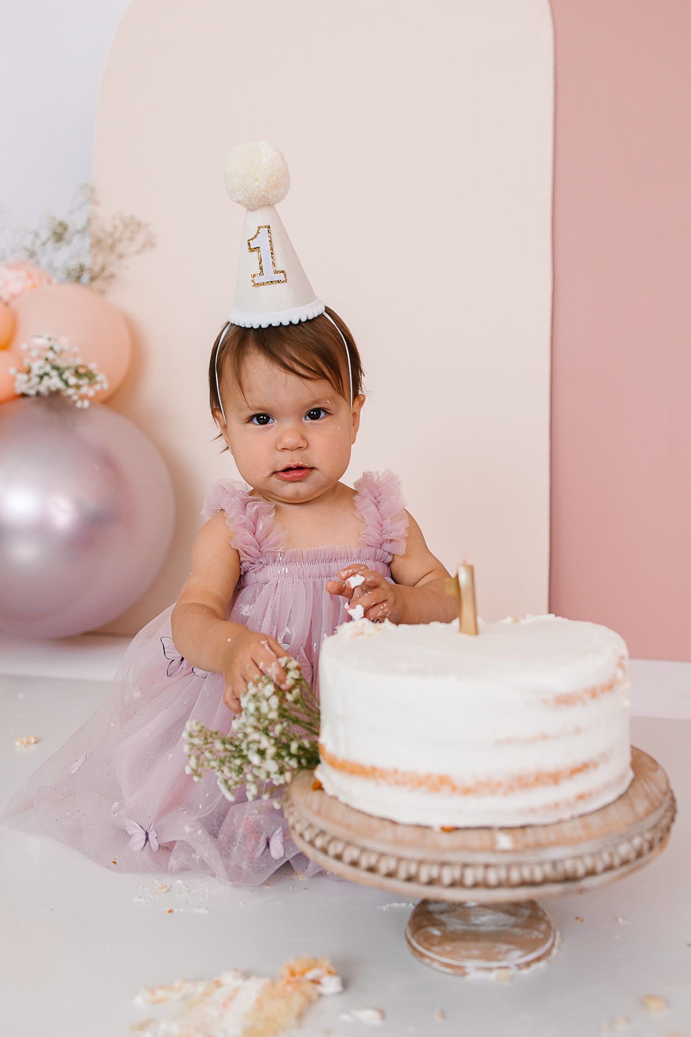 Pink and white first birthday cake smash photo session featuring a delicate cake on a stand with floral accents.