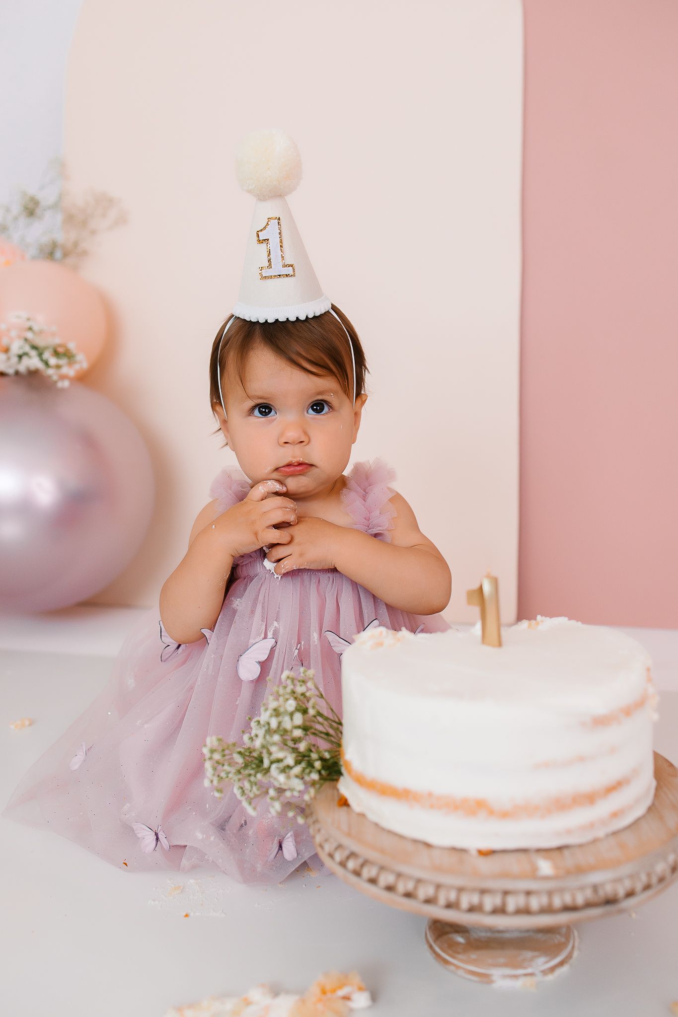 First birthday cake smash photo session featuring a baby girl in a lavender dress with white cake on a stand.