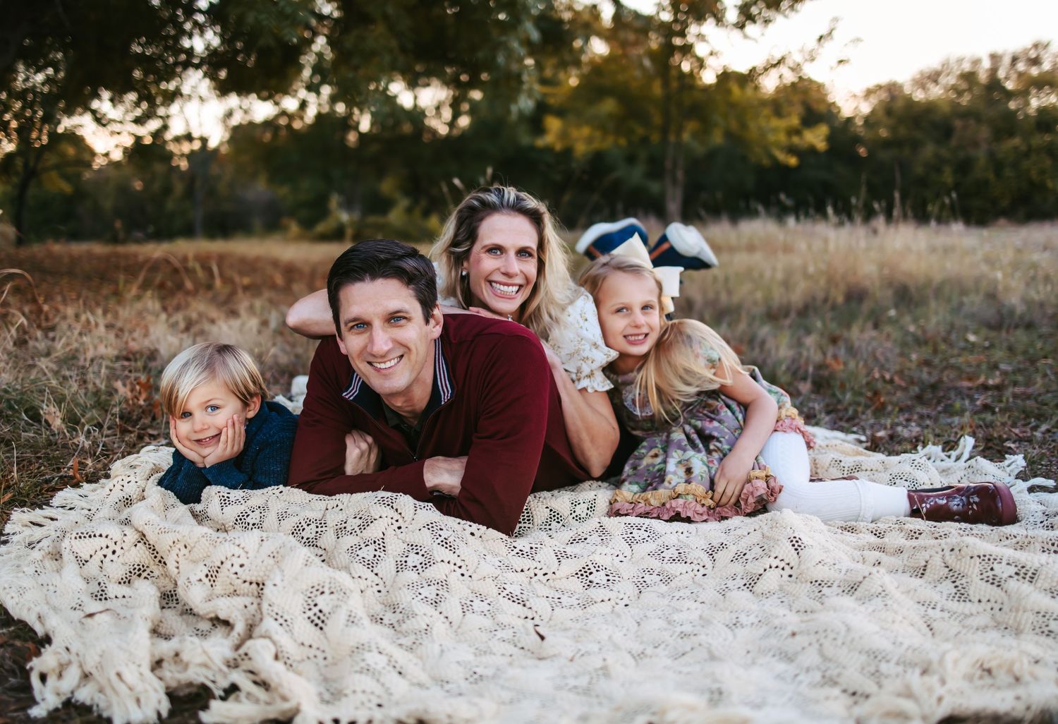 A family lounges together on a cream colored blanket in a rustic outdoor setting during golden hour.