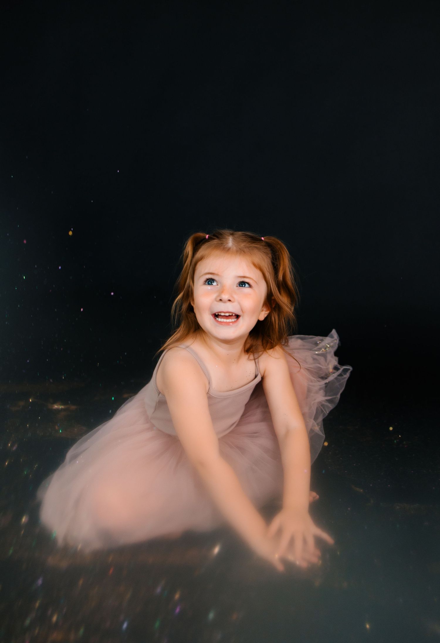 A child in a pink dress lies on a dark floor with glitter and sparkles creating a magical atmosphere.