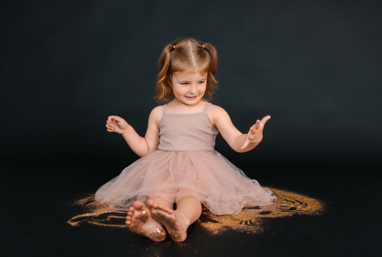 A small child in a pink tulle dress sits gracefully on a dark black backdrop playing in glitter.