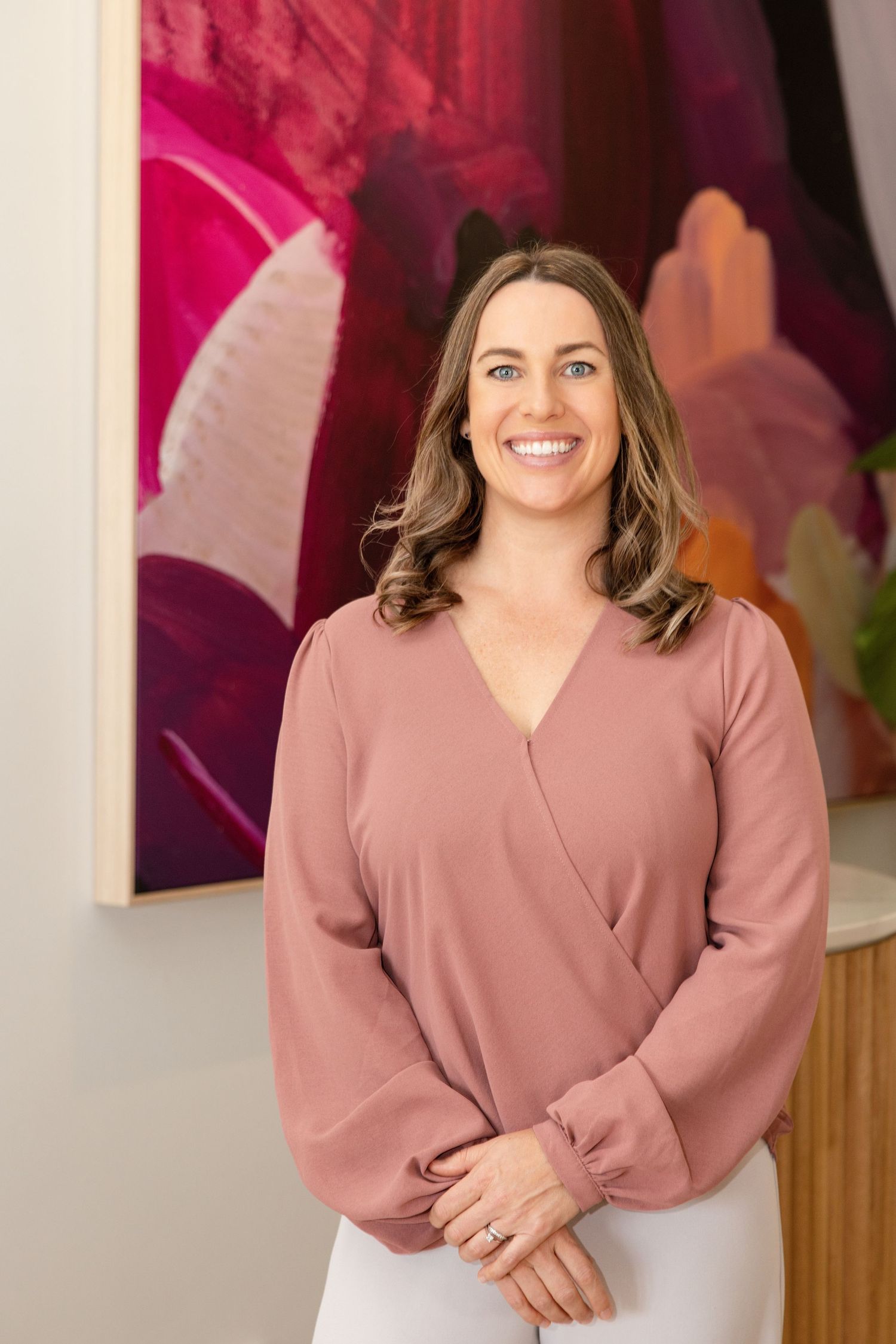 Smiling woman in a pink wrap blouse standing in front of colorful abstract artwork in a modern interior.