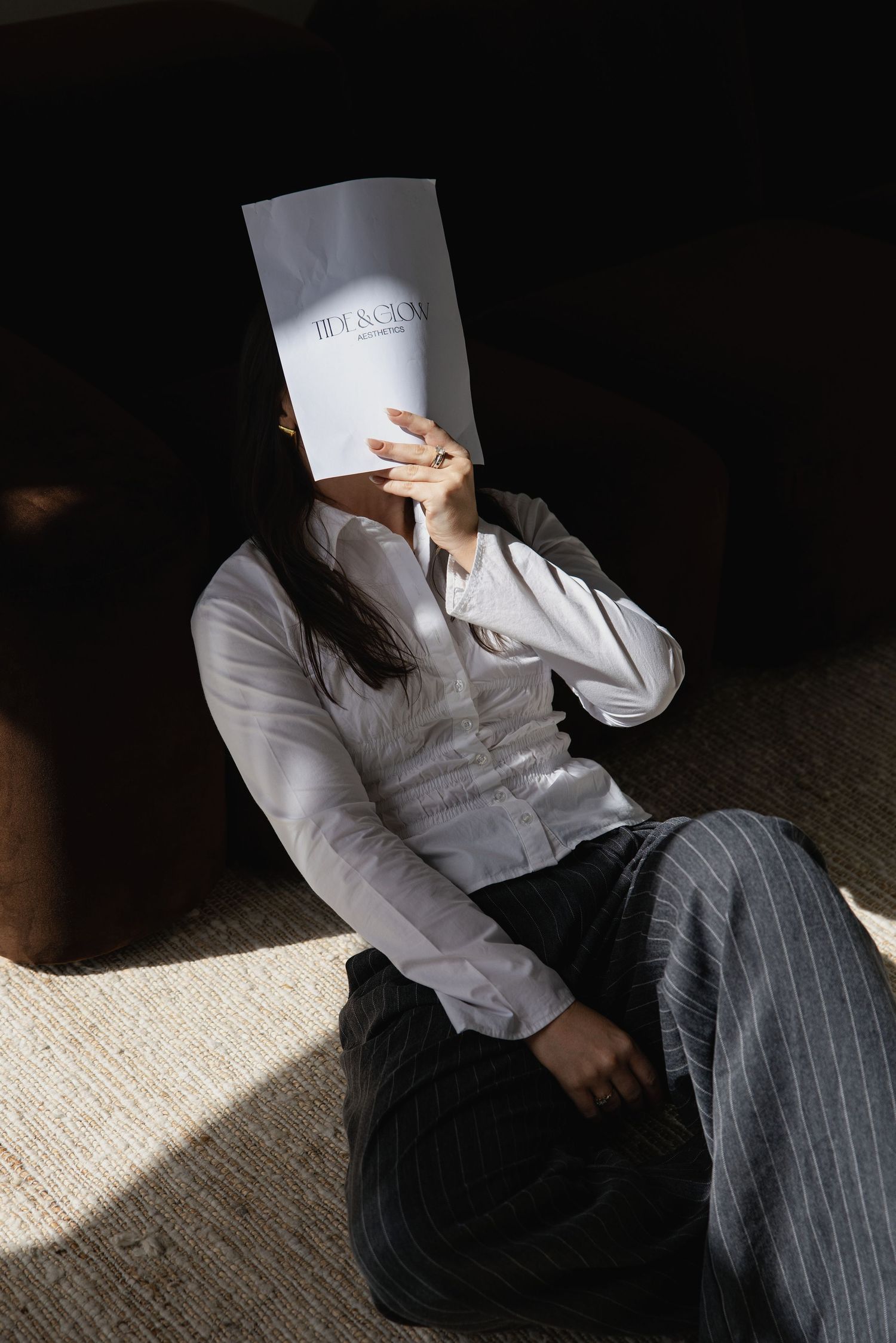 Person in white shirt and striped pants sitting on floor holding paper over face in sunlight.