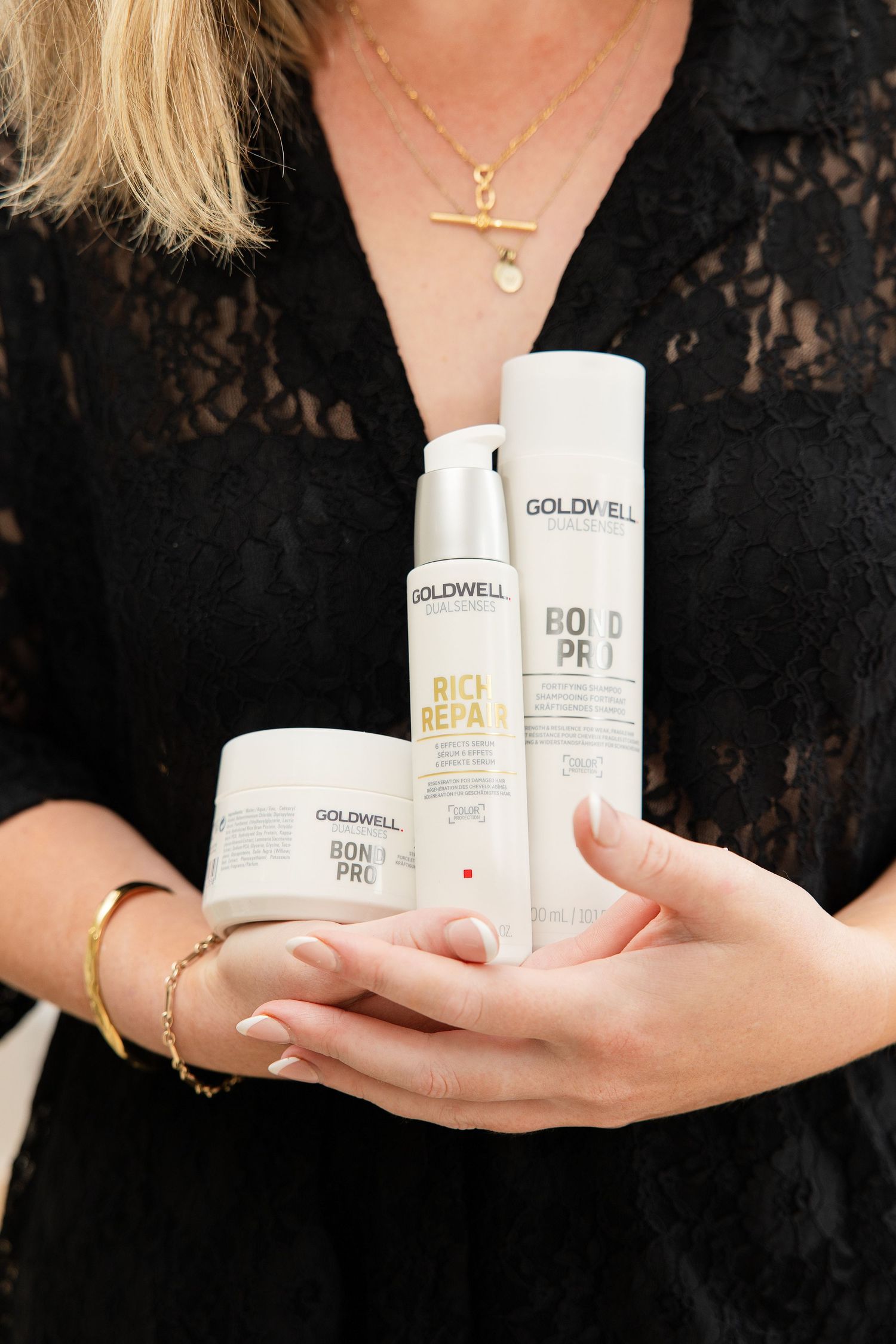 Woman in black lace dress holding three Goldwell Bond Pro hair care products including a serum, mask, and conditioner.