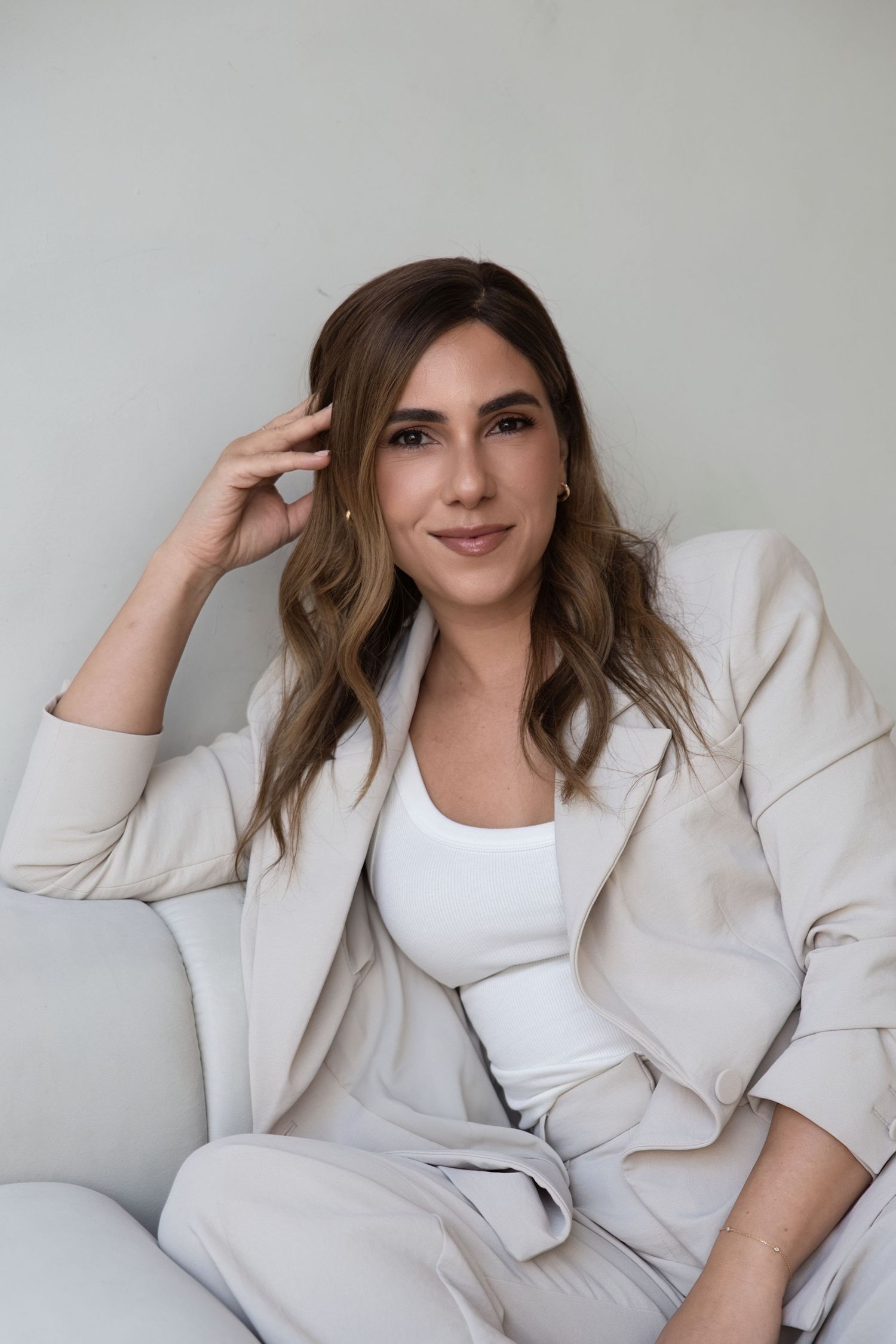 Professional woman in beige blazer suit sitting relaxed, smiling confidently against neutral background.