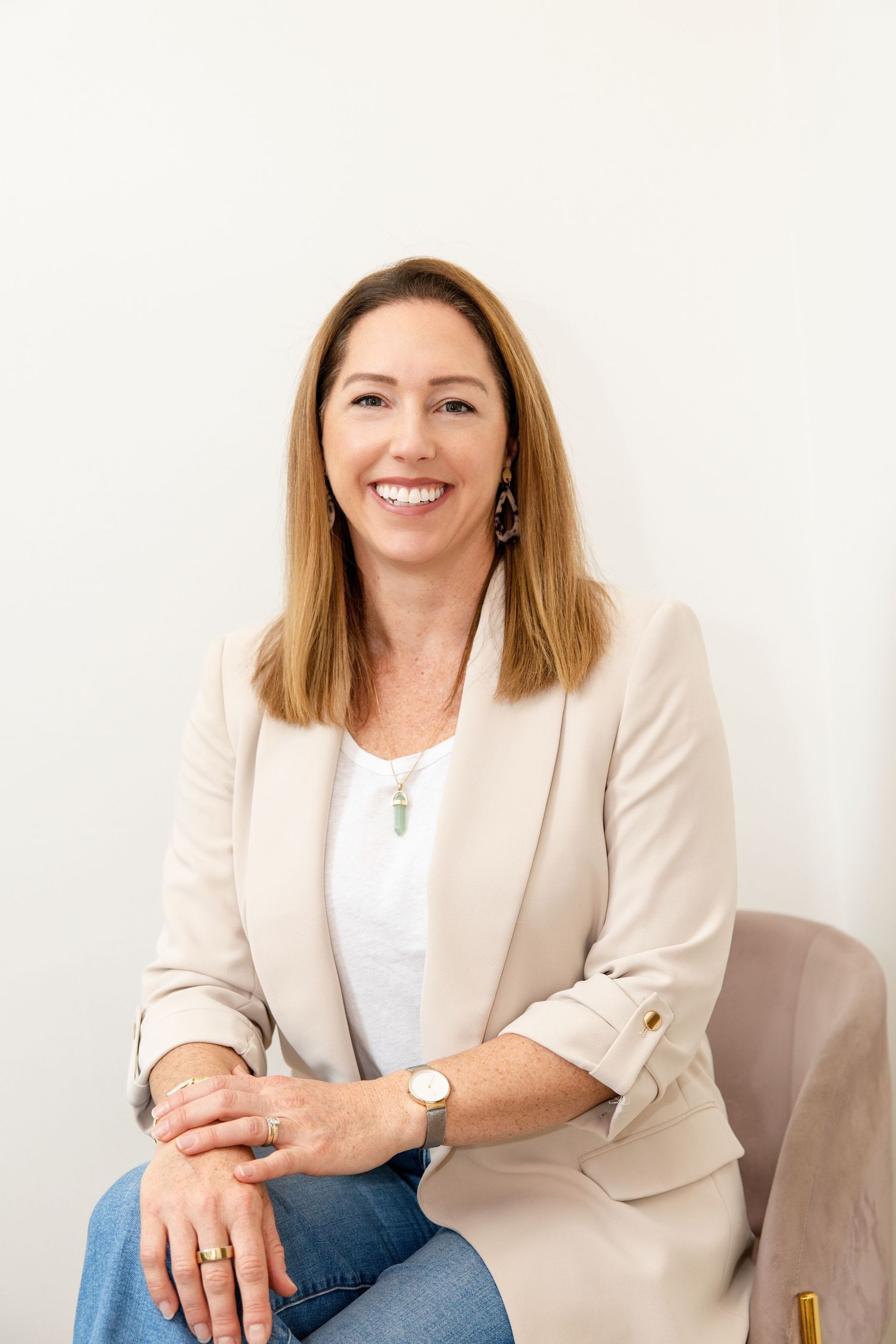 Smiling woman in beige blazer and jeans sitting on chair against white wall wearing crystal pendant necklace.