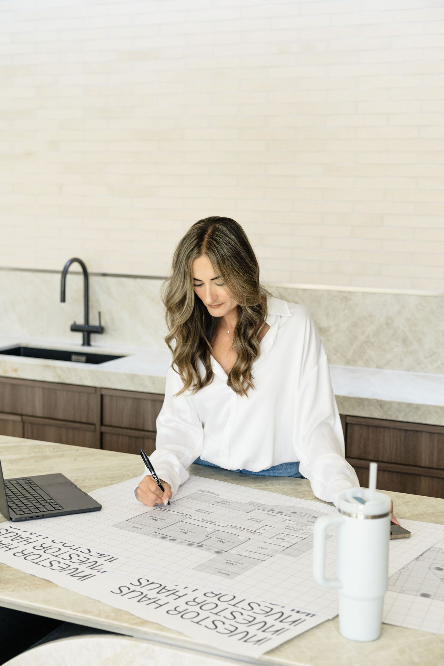 Woman in white shirt reviews floor plans on a kitchen counter beside a laptop and tumbler.
