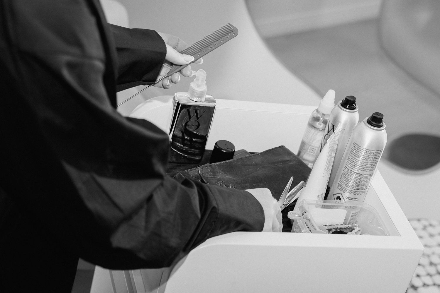 Barber in black gloves selecting styling products from a salon station in a black and white photo.