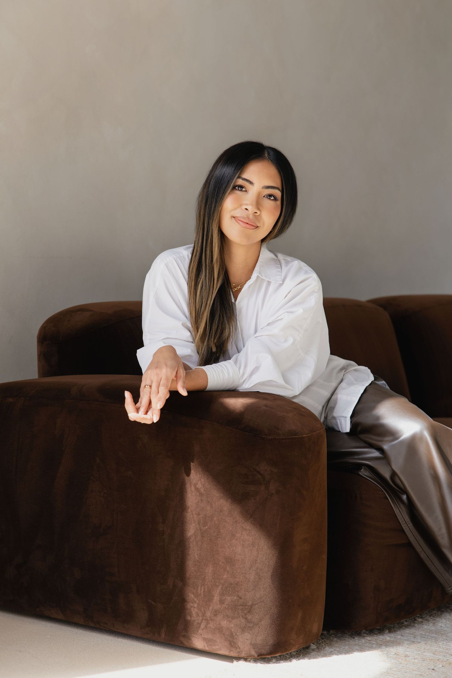 Young woman in white shirt and leather pants relaxing on brown velvet sofa in modern room.