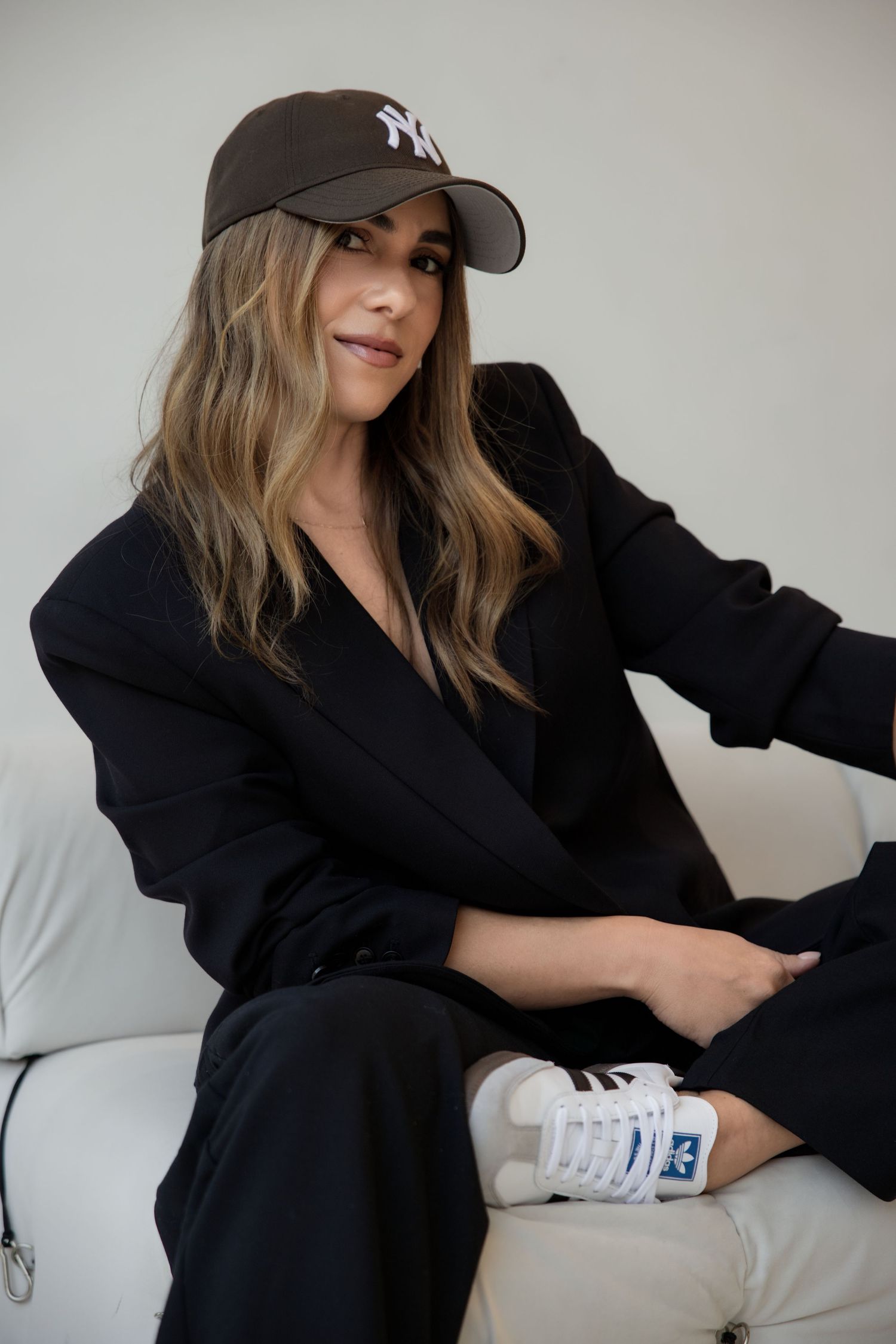 Stylish woman in black suit and NY Yankees cap posing on white sofa wearing white Adidas sneakers.