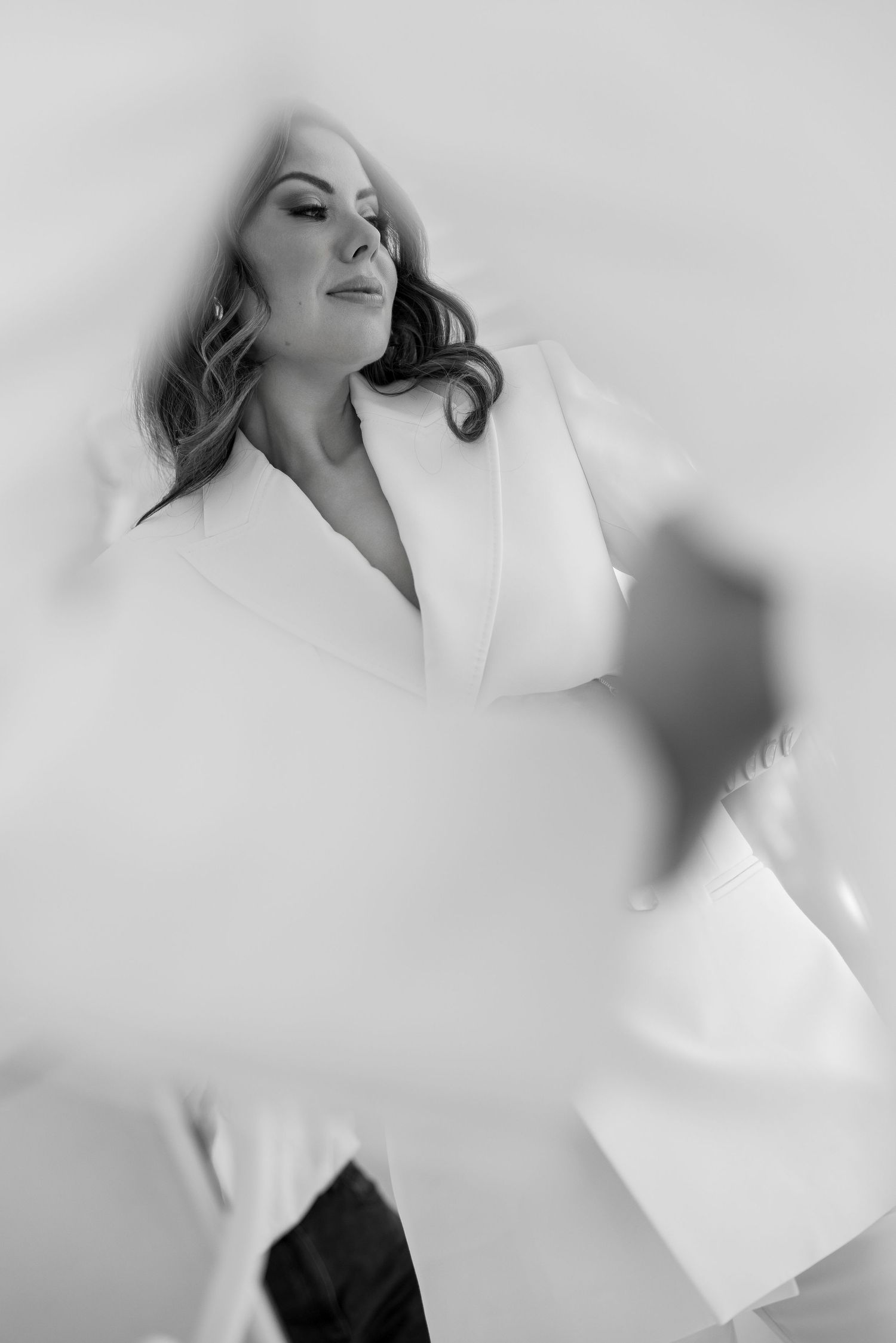 Black and white photo of a woman in a white blazer posing gracefully with soft focus foreground elements.
