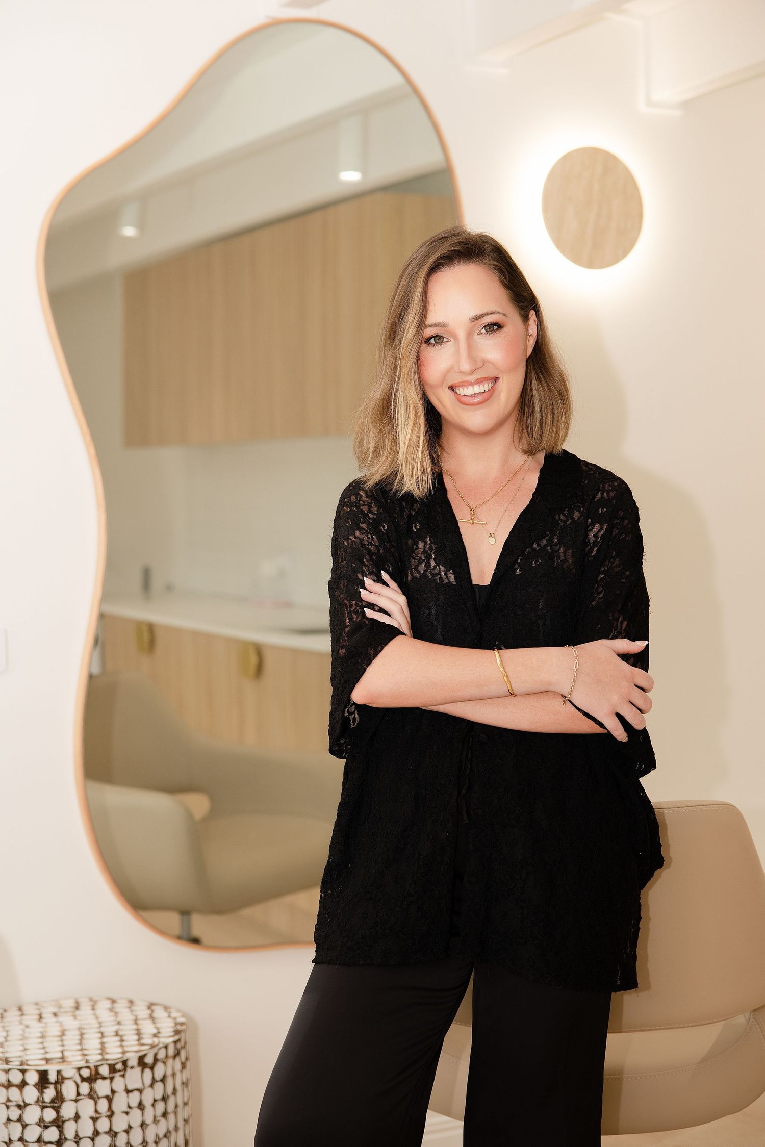 Smiling woman in black lace top with arms crossed stands in modern salon interior with decorative mirror.