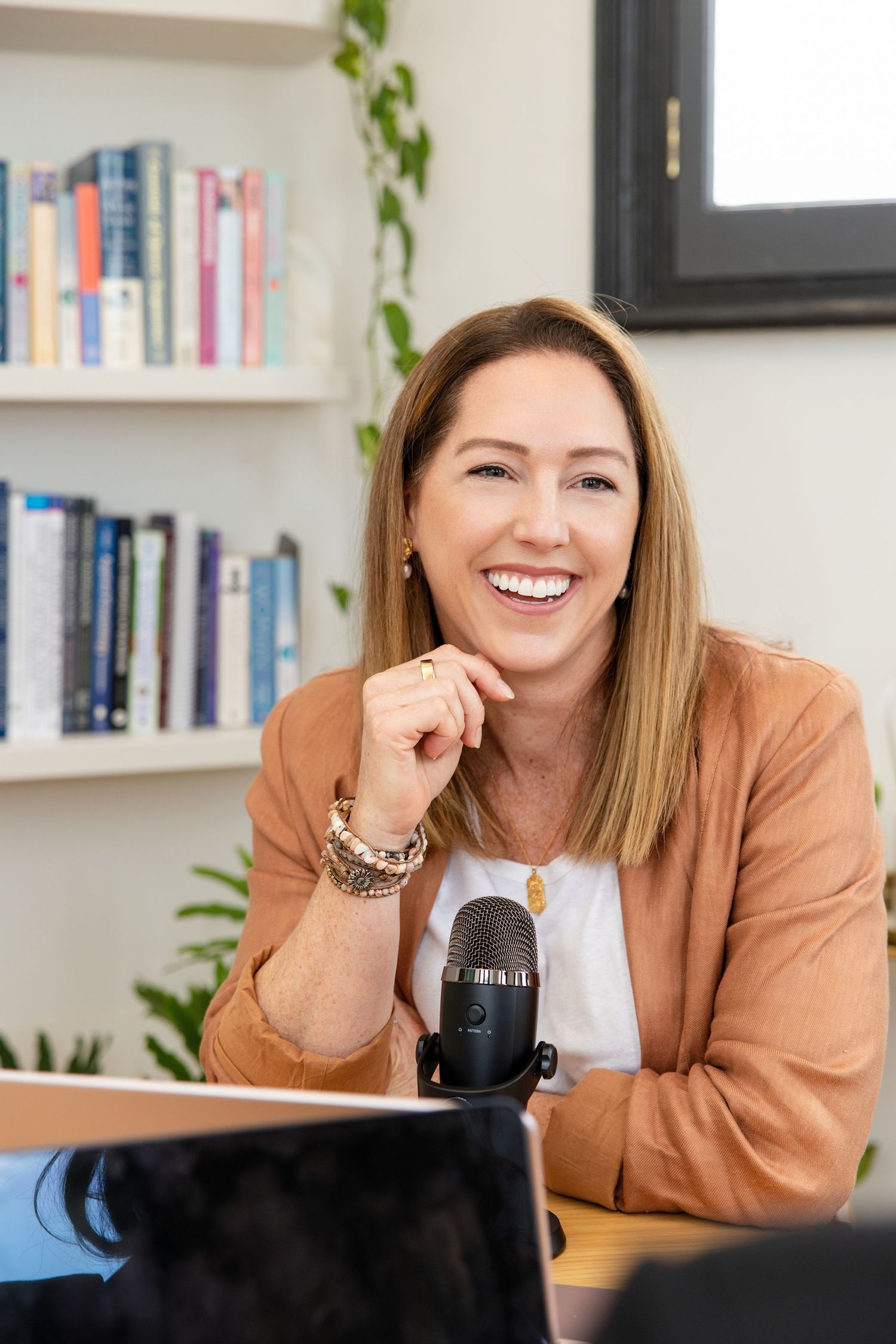 A laughing blonde woman in a tan blazer sitting at a desk with a podcast microphone in a bright office.