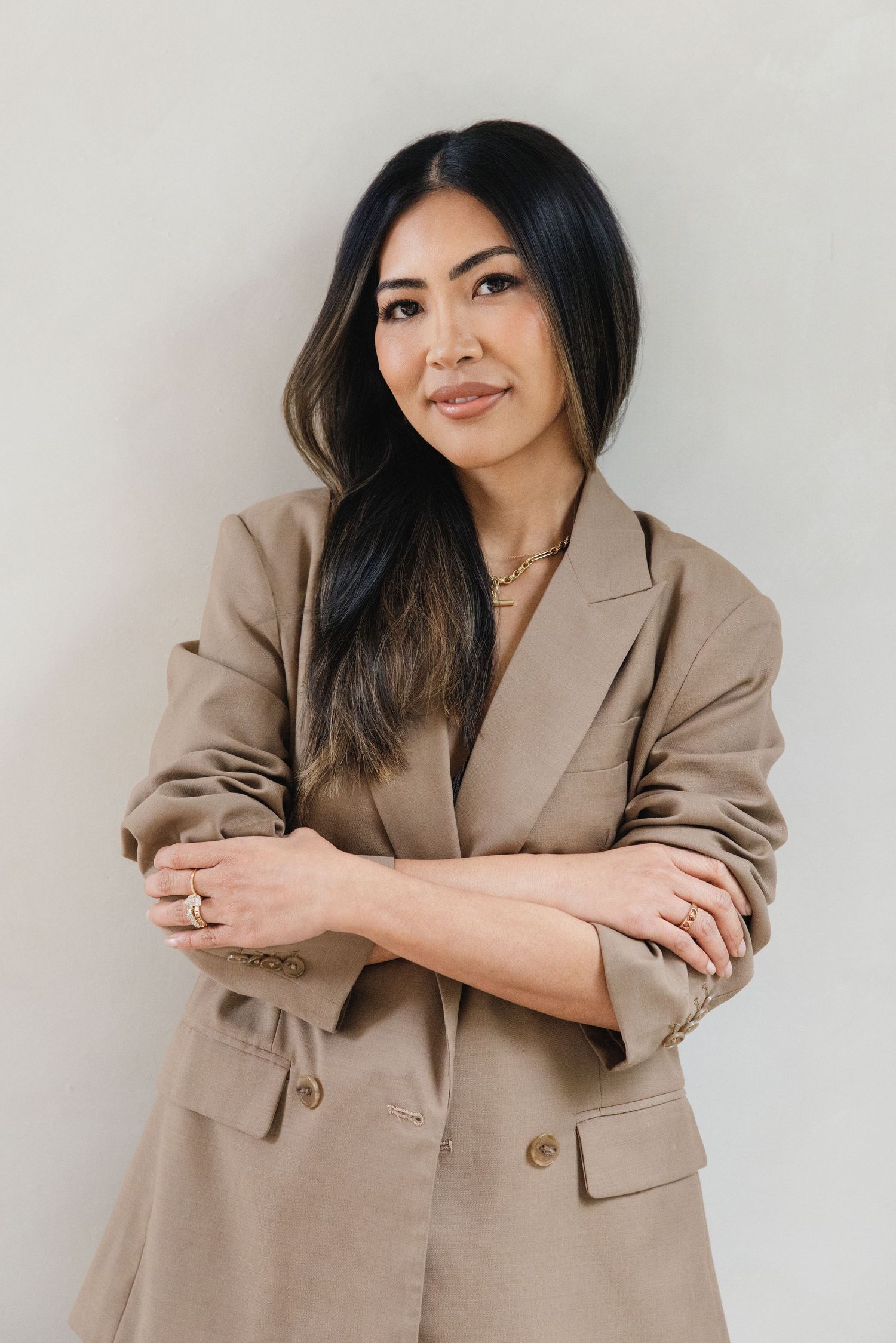 Professional Asian woman in beige blazer posing confidently with arms crossed against neutral background.