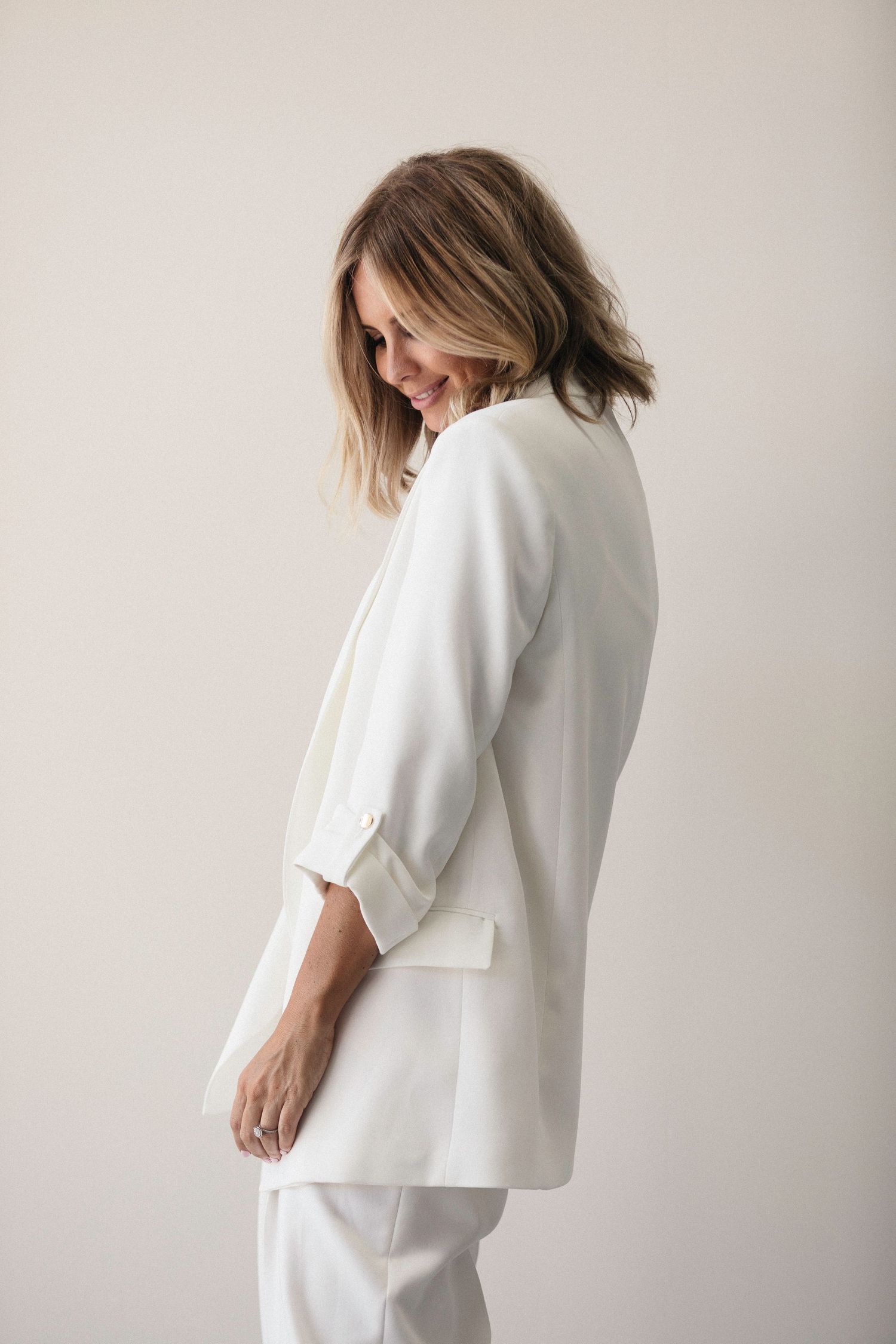Woman smiling in a chic white blazer and pants suit, posed sideways against a light background.