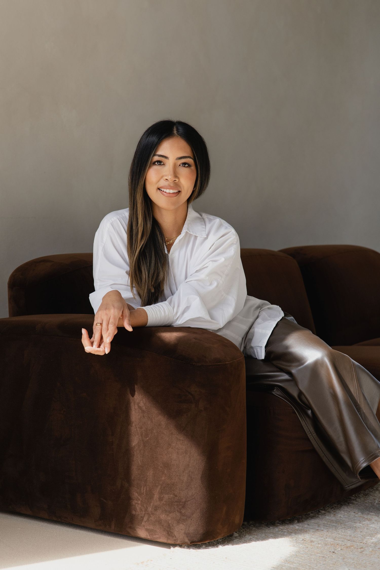 Smiling woman in white shirt seated on brown velvet couch with sunlight in neutral interior.