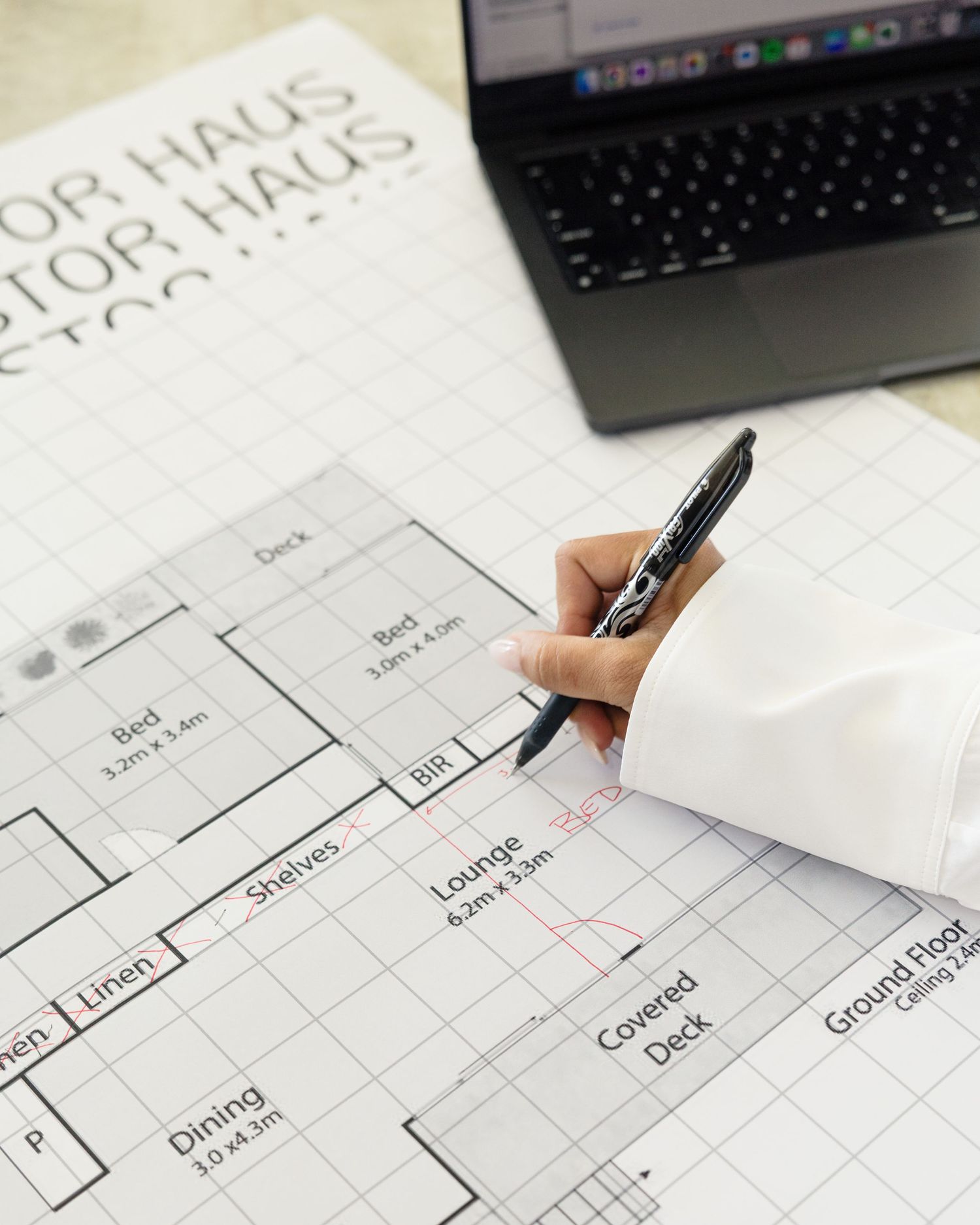 Hand marking architectural floor plans on grid paper beside a laptop on a desk.