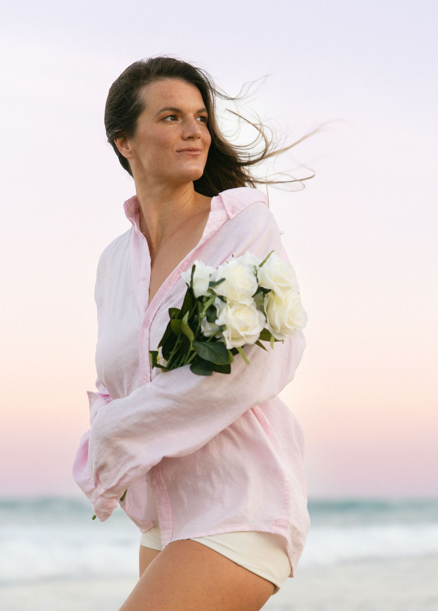 A breezy beach scene with white roses and flowing fabric against a sunset sky and ocean waves.