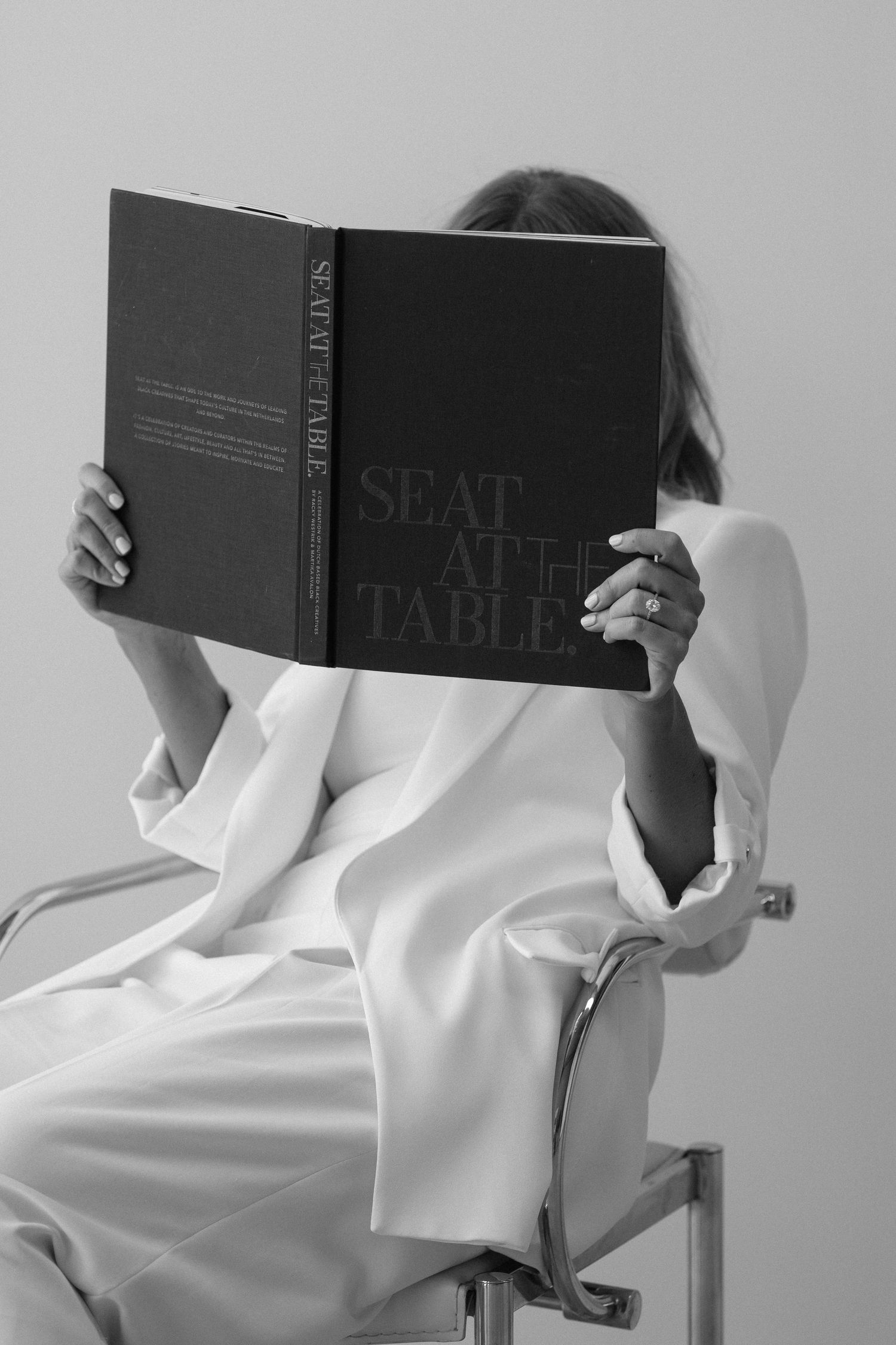Woman in white blazer reading a dark hardcover book titled Seat at the Table while seated on a modern chair.