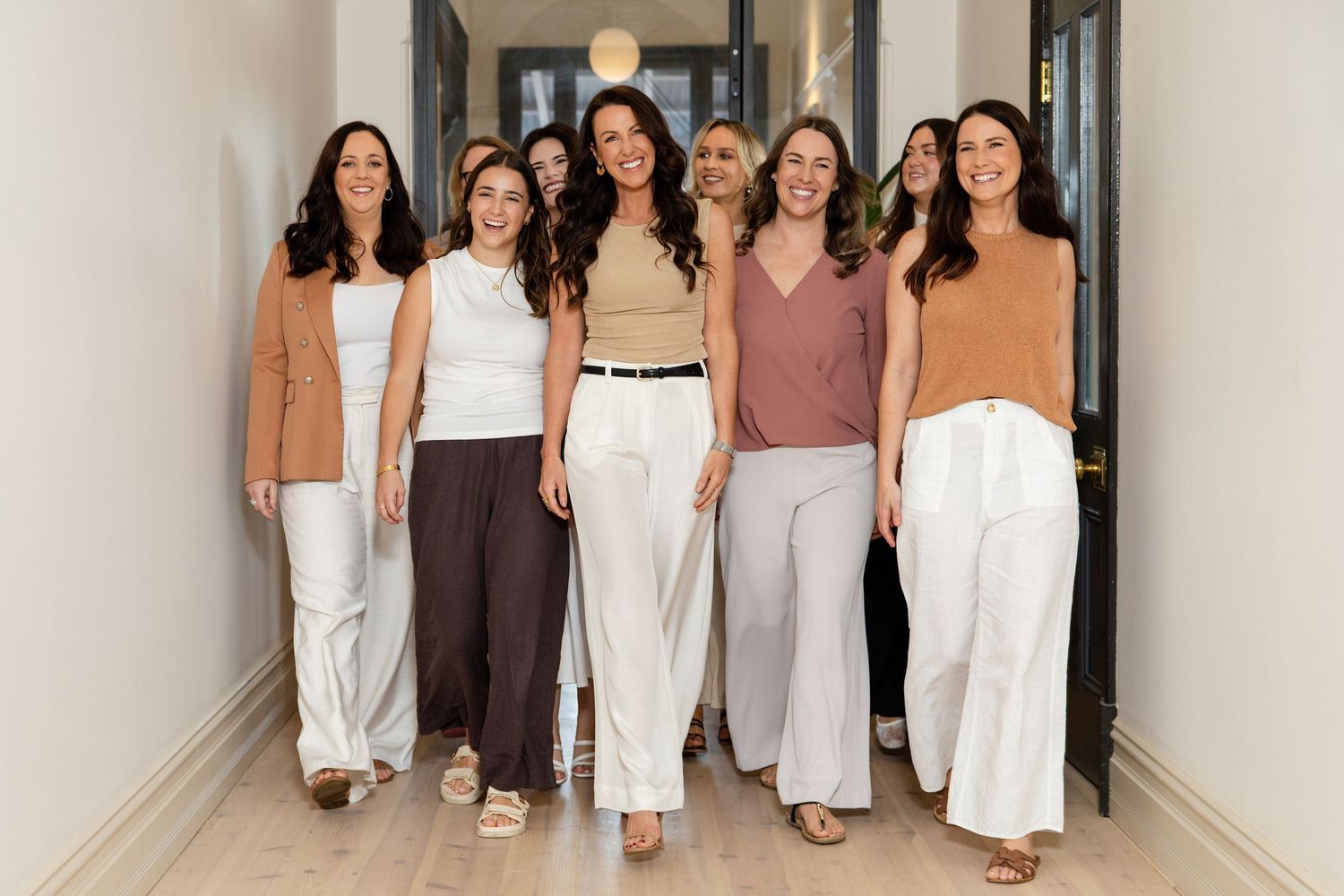 A group of eight women in neutral tones walking confidently down a bright hallway.