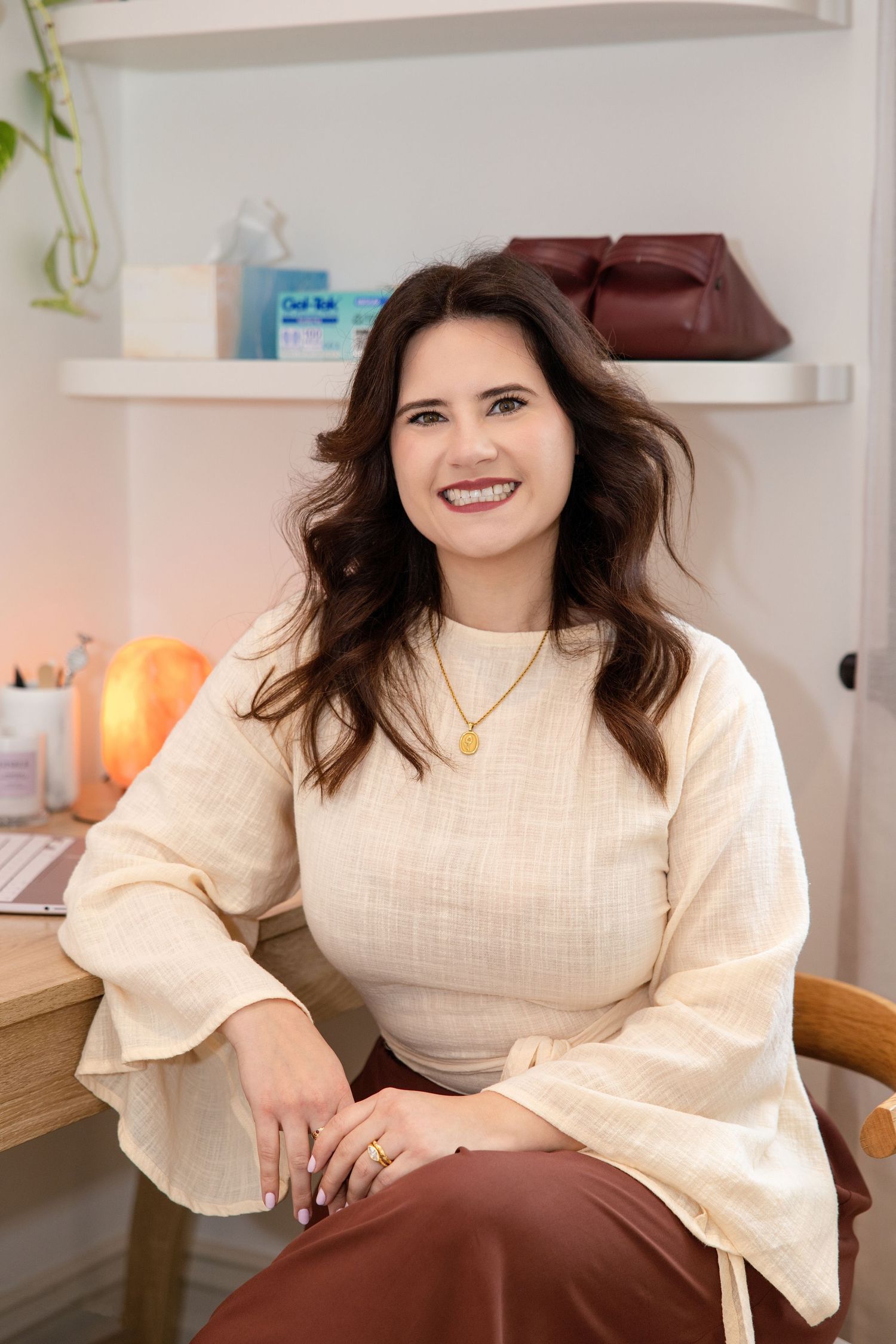 Smiling brunette woman in cream blouse and burgundy skirt sitting in bright, modern office space with shelf decor.