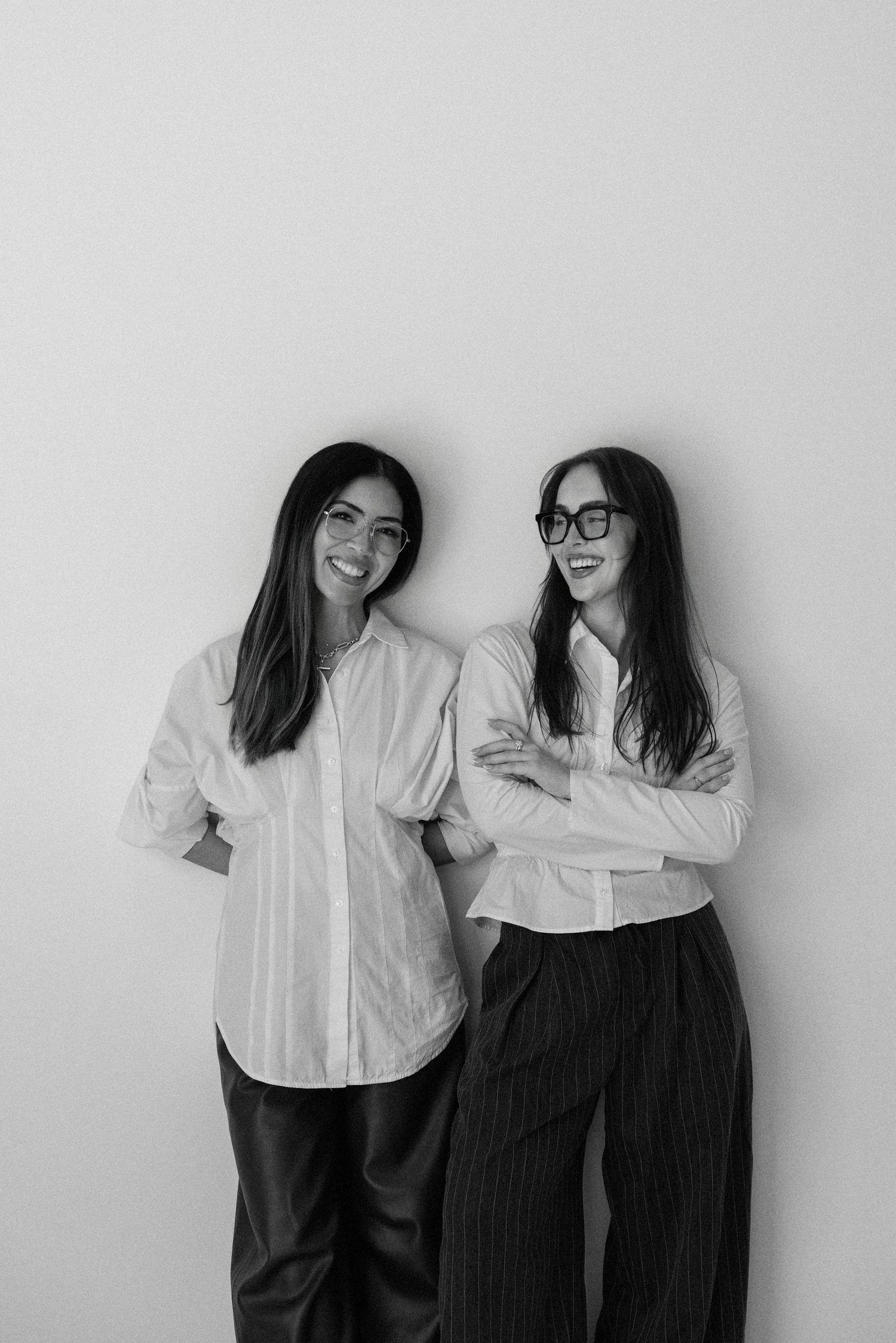 Two smiling women in white button-up shirts pose together against a light background in black and white photo.