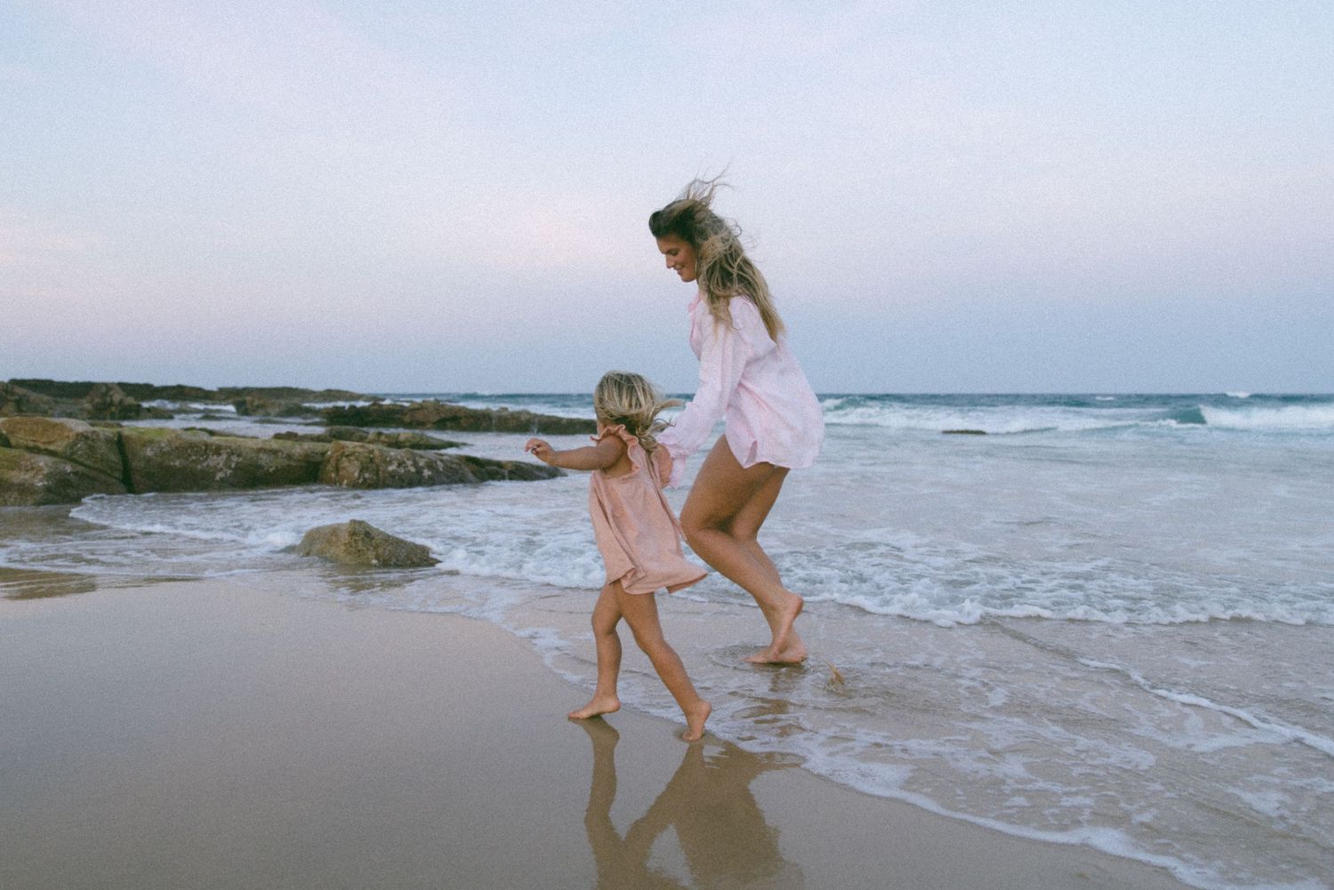 Two people walk along a serene beach at sunset while waves gently roll onto the shore.