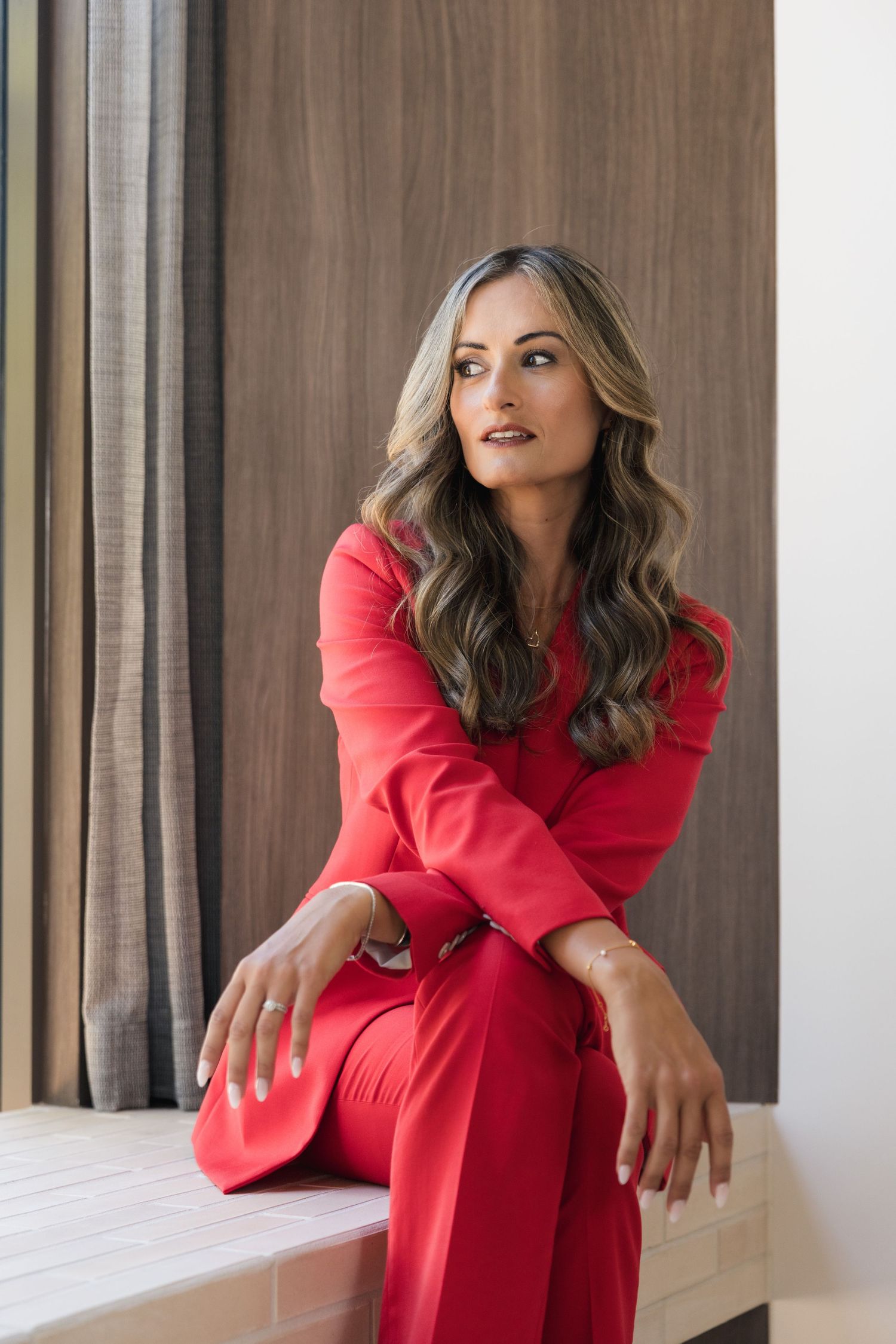 Brunette woman in red blazer suit sitting confidently near curtained window indoors.