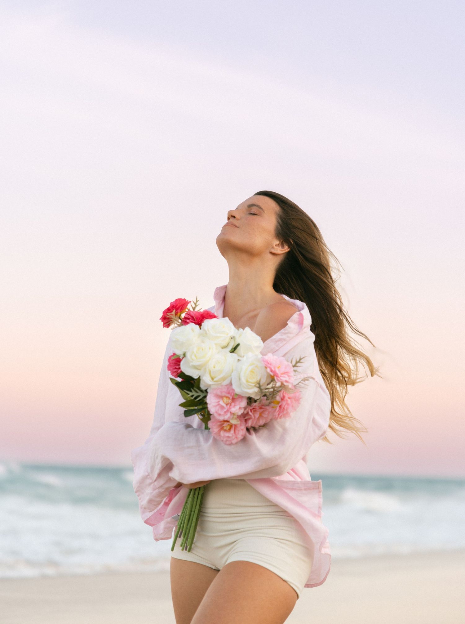 Person in white outfit dancing with pink flowers on beach during sunset.