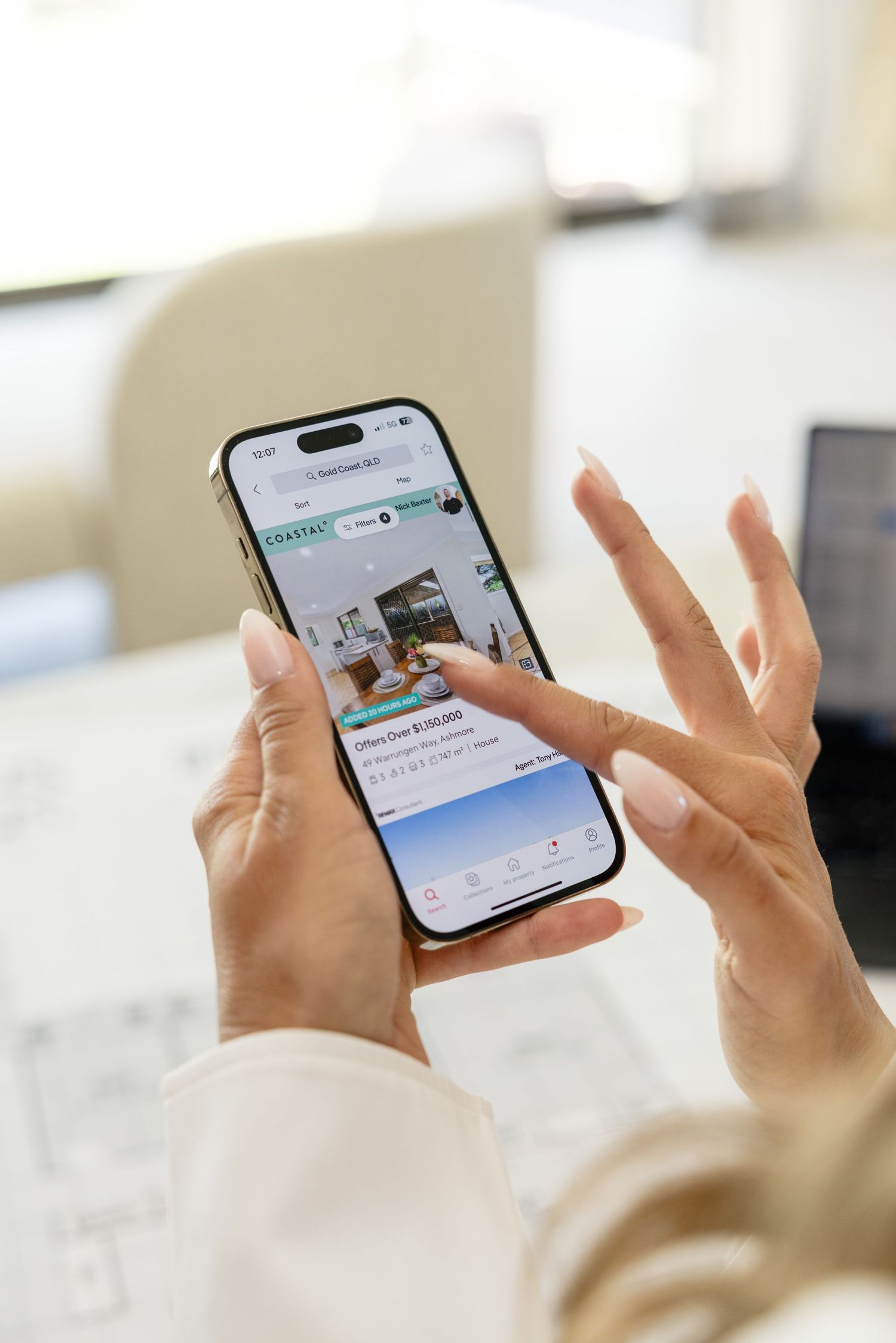 Woman browsing a real estate app on iPhone, scrolling through property listings at a desk.
